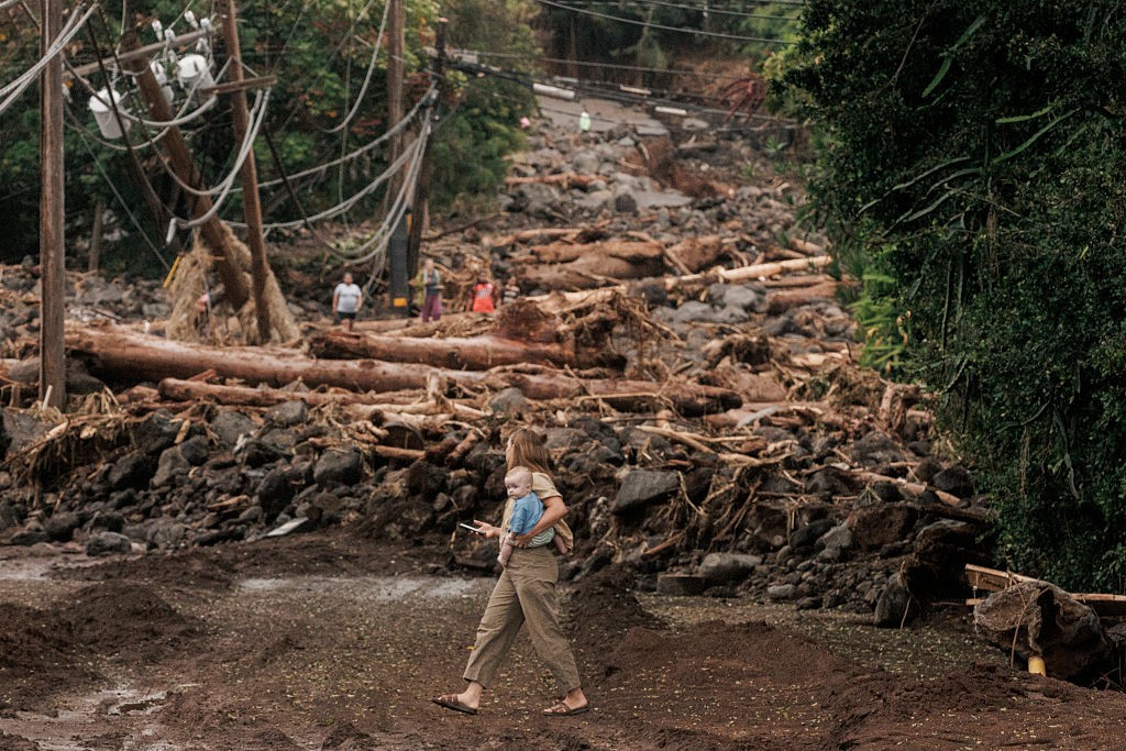 Mulher com bebê no colo caminha por estrada de terra e lama, com postes de energia caídos e destroços de árvores e pedras ao fundo, após um desastre natural. Outras pessoas observam a devastação.