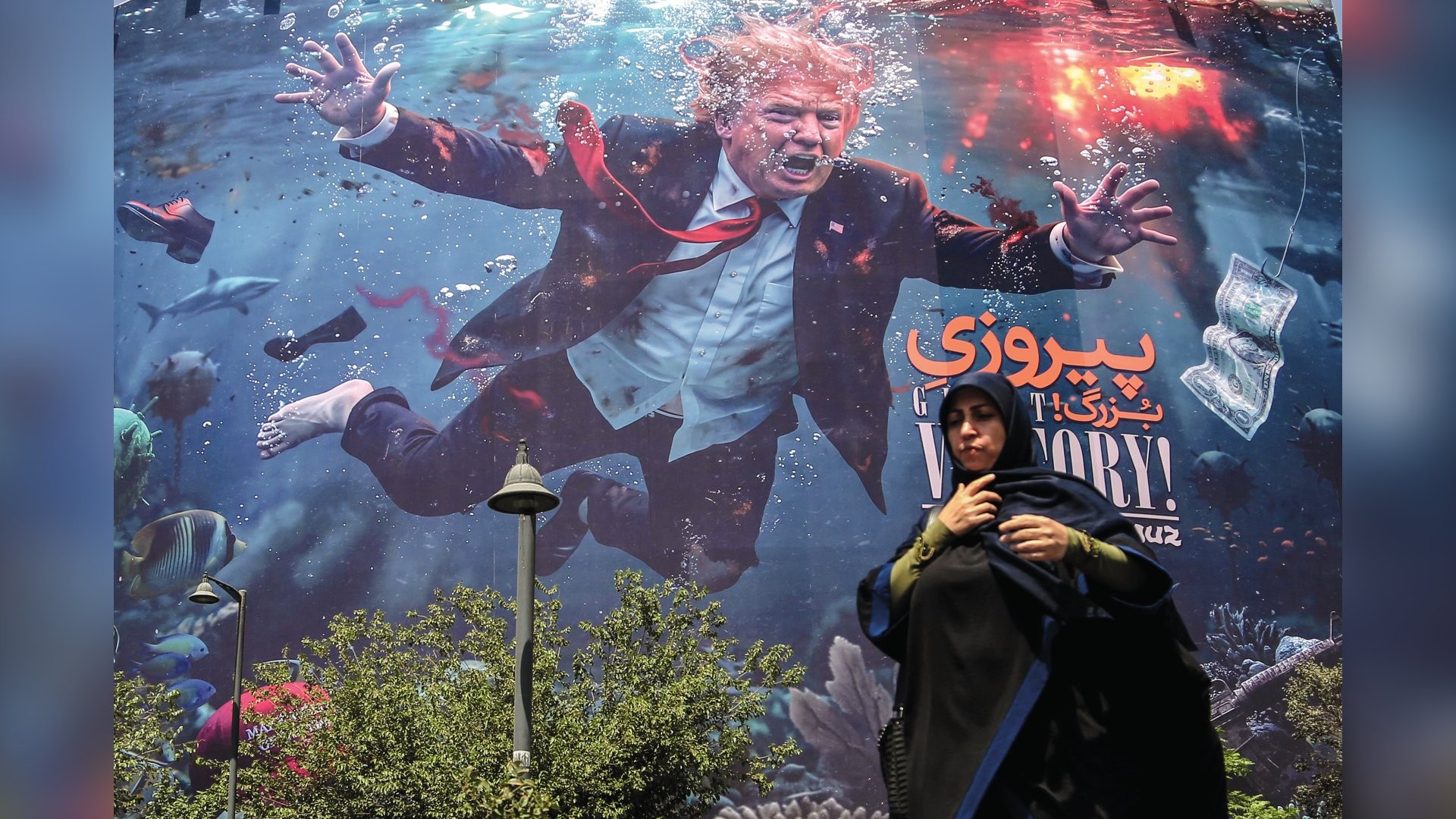 Mulher de hijab preto e blusa verde-oliva em primeiro plano, com um outdoor ao fundo. O outdoor mostra Donald Trump submerso, com a boca aberta, braços estendidos e sapatos flutuando. Peixes, tubarões e notas de dólar em um anzol completam a cena subaquática. Há texto em persa e inglês VICTORY! no outdoor. Arbustos e um poste de luz aparecem na parte inferior da imagem