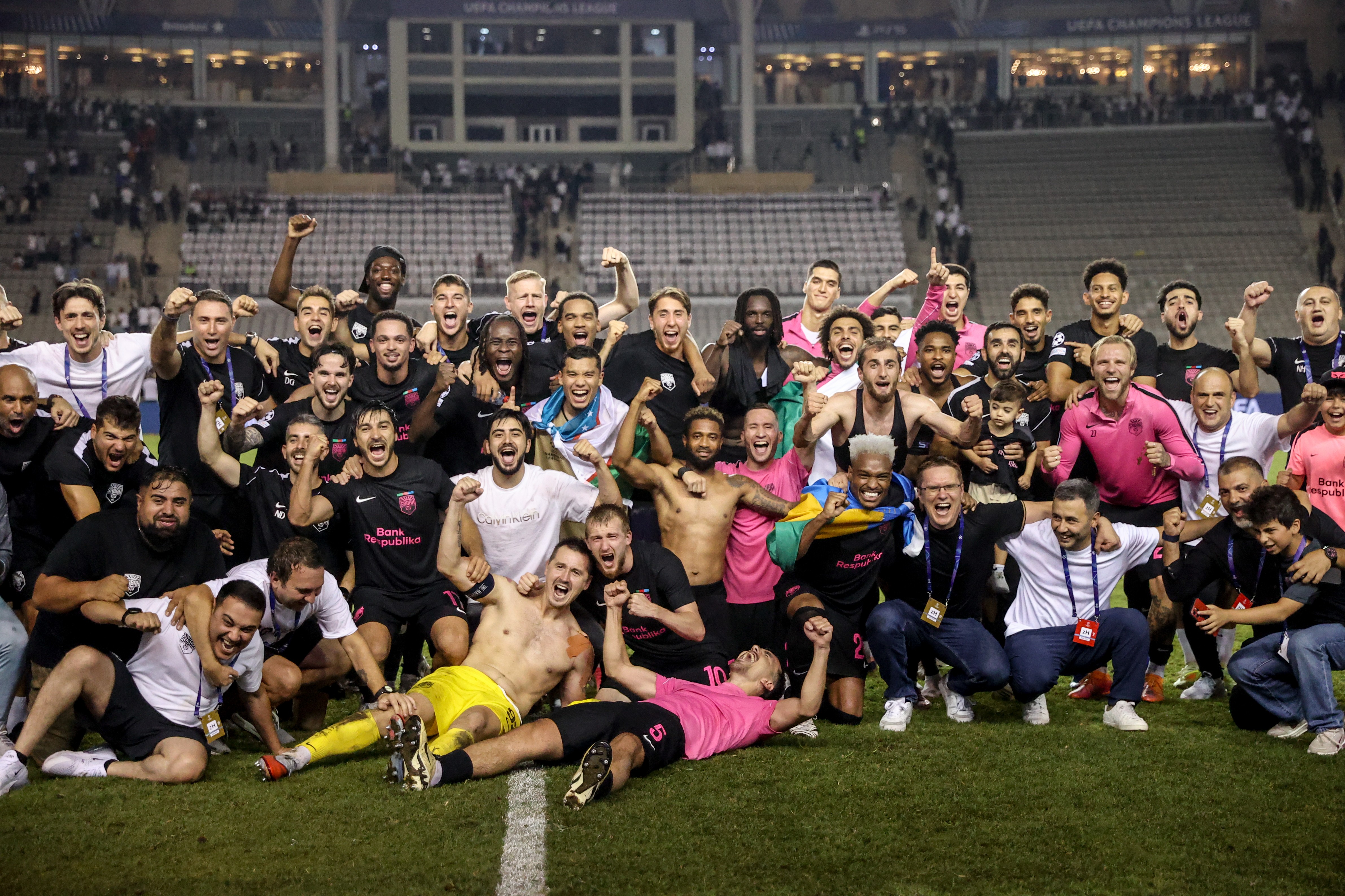Um grupo de jogadores de futebol e membros da comiss&atilde;o t&eacute;cnica, a maioria homens, celebra com euforia no gramado de um est&aacute;dio lotado. Eles sorriem, gritam e levantam os bra&ccedil;os em sinal de vit&oacute;ria. Alguns vestem camisas de futebol pretas ou rosas, outros est&atilde;o sem camisa, e um jogador deitado no ch&atilde;o usa shorts amarelos. Ao fundo, as arquibancadas est&atilde;o cheias de torcedores
