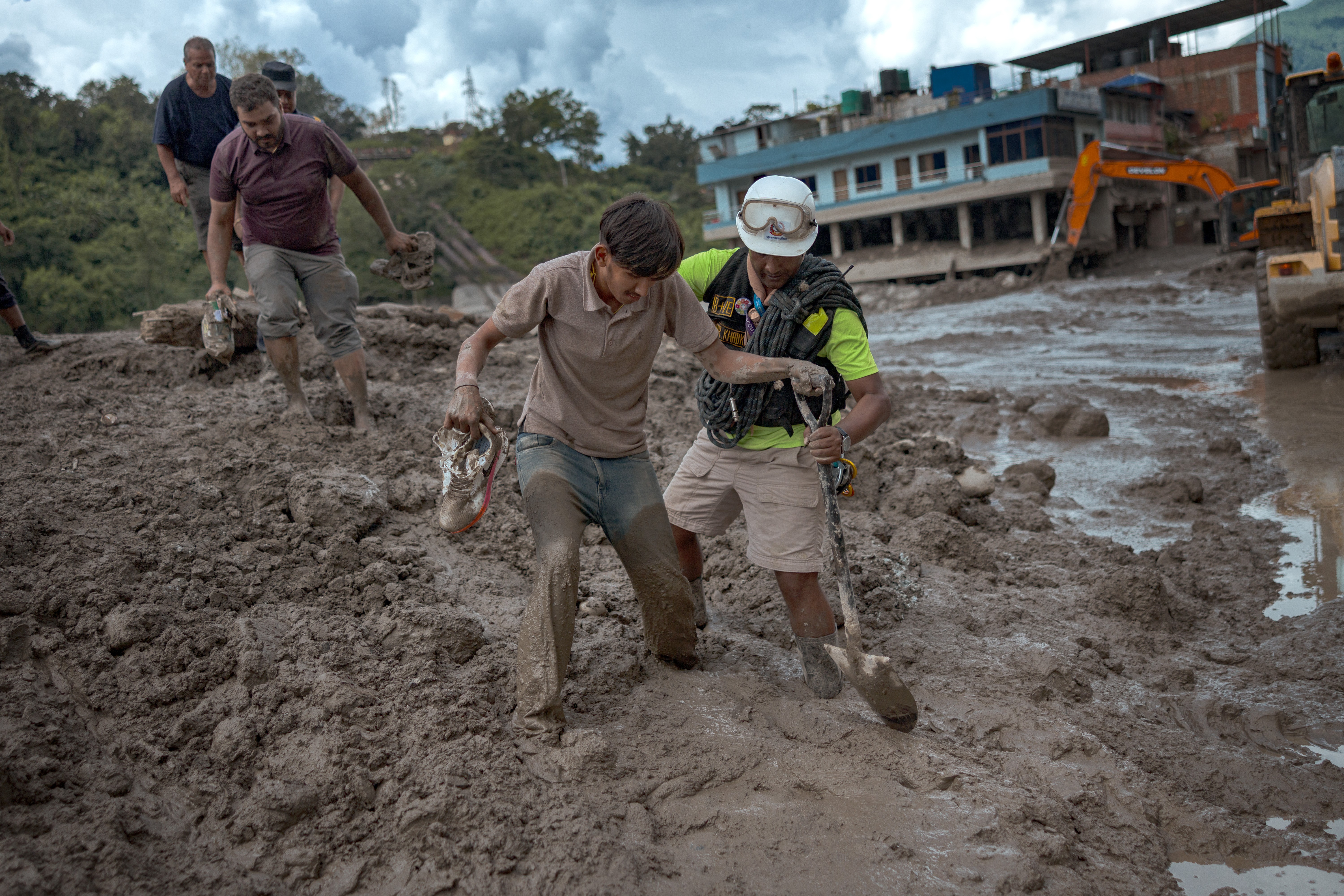 Homens caminham por uma &aacute;rea devastada por lama e detritos. Um homem de camisa polo marrom e cal&ccedil;a jeans suja segura sapatos, enquanto outro, com capacete branco, &oacute;culos e colete de seguran&ccedil;a, carrega uma p&aacute;. Ao fundo, casas danificadas e m&aacute;quinas pesadas em meio &agrave; destrui&ccedil;&atilde;o. Outros homens tamb&eacute;m se movem pela lama. O c&eacute;u est&aacute; nublado, indicando um cen&aacute;rio p&oacute;s-desastre.