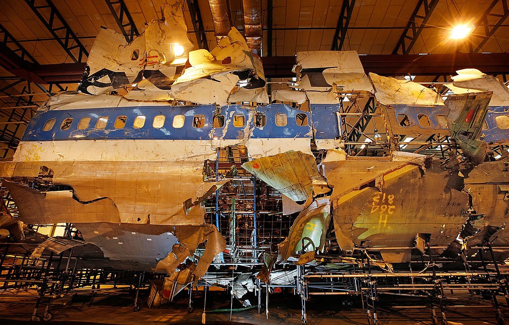 Destro&ccedil;os de um avi&atilde;o com fuselagem azul e branca, rasgada em v&aacute;rias partes, exp&otilde;em a estrutura interna met&aacute;lica. As janelas est&atilde;o vis&iacute;veis na parte superior, e as pe&ccedil;as est&atilde;o montadas sobre andaimes em um galp&atilde;o com ilumina&ccedil;&atilde;o quente