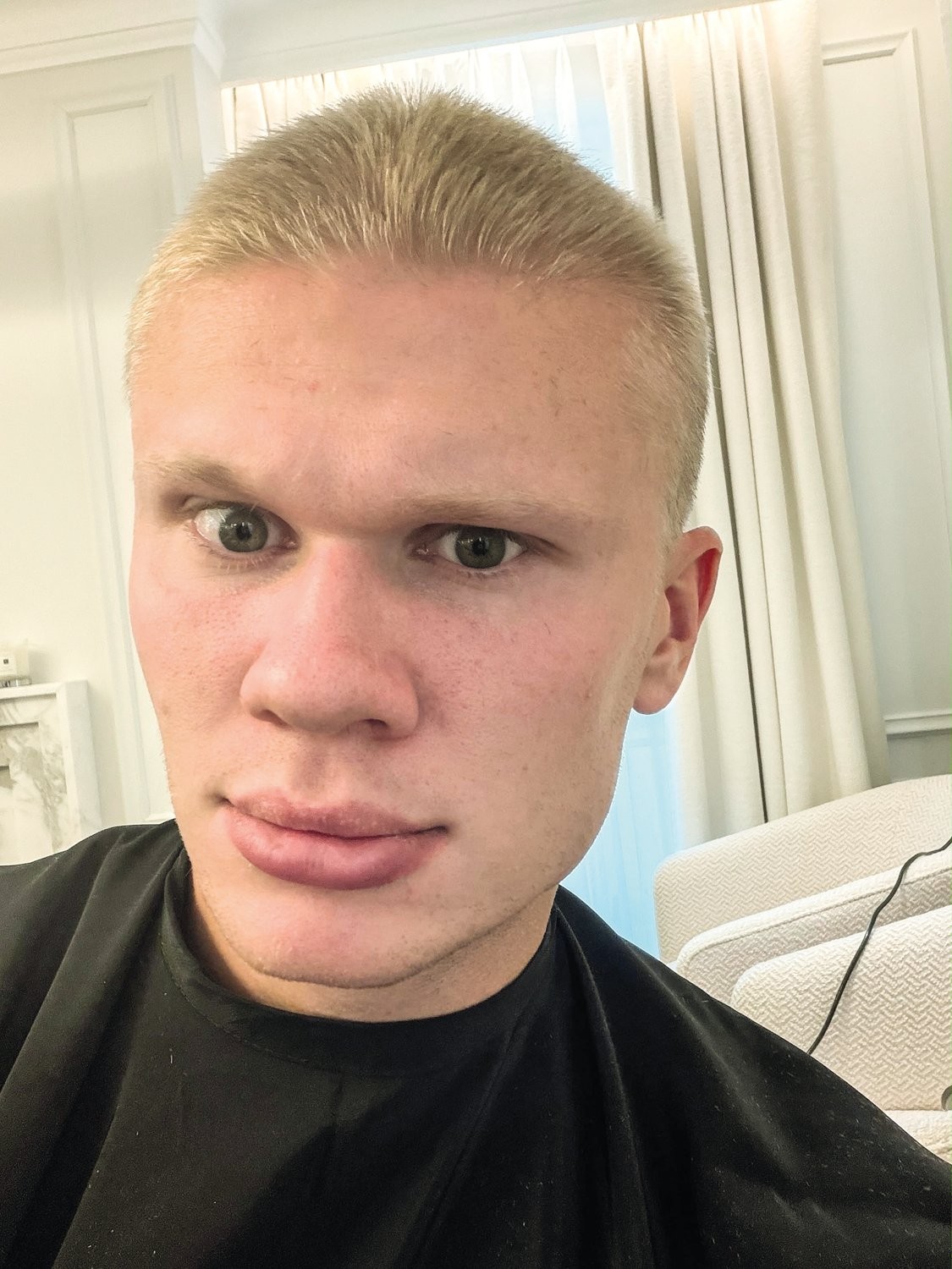 Rosto de Erling Haaland, jogador de futebol, com cabelo loiro curto e olhos verdes, usando uma capa preta de cabeleireiro, olhando diretamente para a c&acirc;mera com uma express&atilde;o s&eacute;ria. Ao fundo, cortinas claras e um sof&aacute; bege.