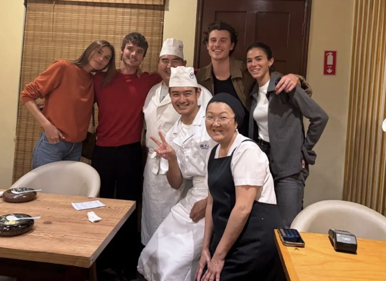 Sete pessoas sorridentes posam em um restaurante. Quatro clientes, incluindo Shawn Mendes e Bruna Marquezine, abraçam três chefs asiáticos, dois homens e uma mulher, que vestem uniformes brancos e pretos. Uma mesa de madeira com pratos e celulares está em primeiro plano