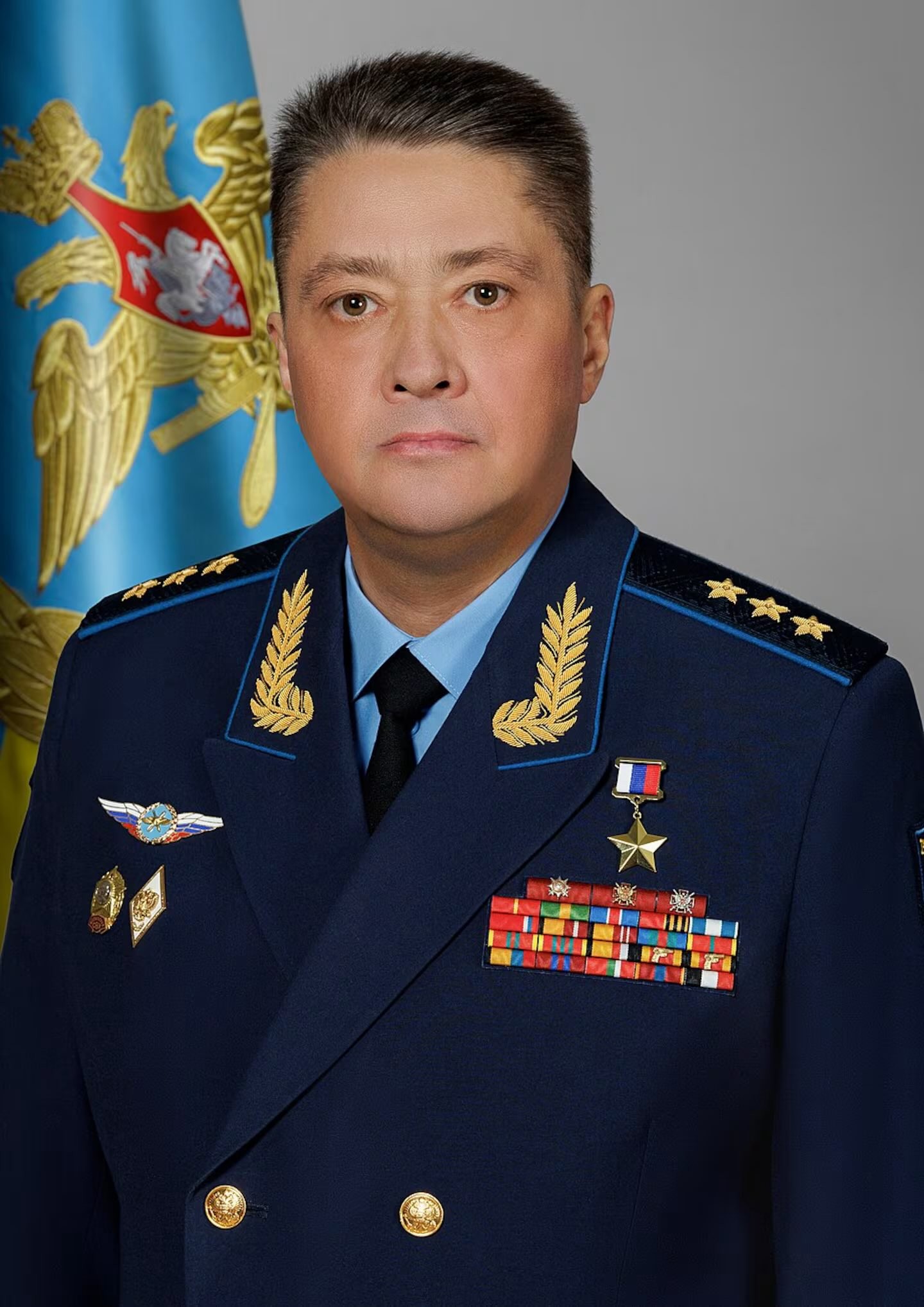 Homem de meia-idade, pele clara e cabelo escuro, vestindo um uniforme militar azul-marinho com insígnias douradas nos ombros e lapelas, gravata preta e camisa azul-clara. Ele exibe diversas medalhas e fitas coloridas no peito, com uma bandeira azul e dourada ao fundo. Sua expressão é séria e ele olha diretamente para frente