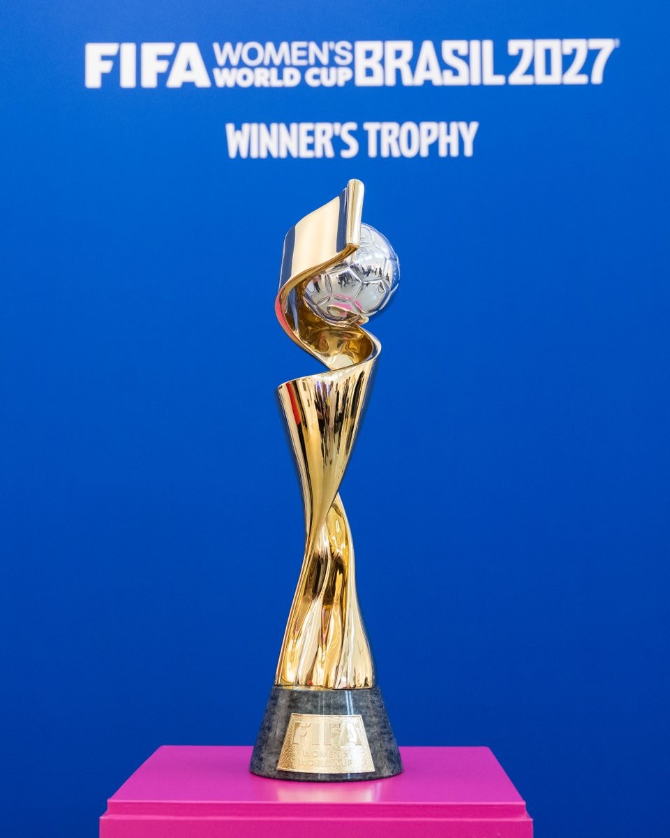 Trof&eacute;u dourado da Copa do Mundo Feminina da FIFA, com uma bola de futebol prateada no topo, sobre uma base escura. Ao fundo, um painel azul com os dizeres FIFA Womens World Cup Brasil 2027 Winners Trophy em branco