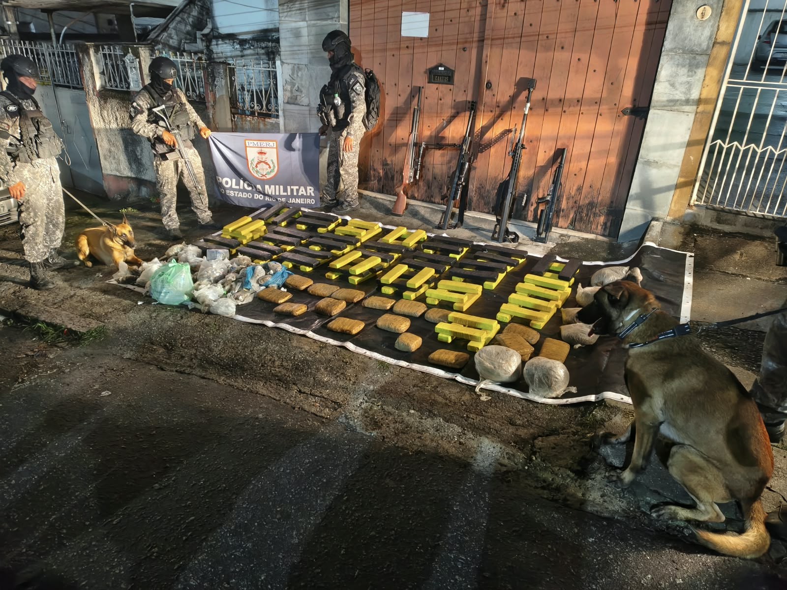 Três policiais militares fardados, com cães farejadores, exibem grande quantidade de drogas e armas apreendidas em uma rua. No chão, sobre um pano branco, estão dezenas de tabletes de substâncias ilícitas, alguns amarelos e outros marrons, além de pacotes menores. Atrás, encostadas em uma porta de madeira, há quatro armas de fogo longas. Um banner da Polícia Militar do Estado do Rio de Janeiro está estendido no centro