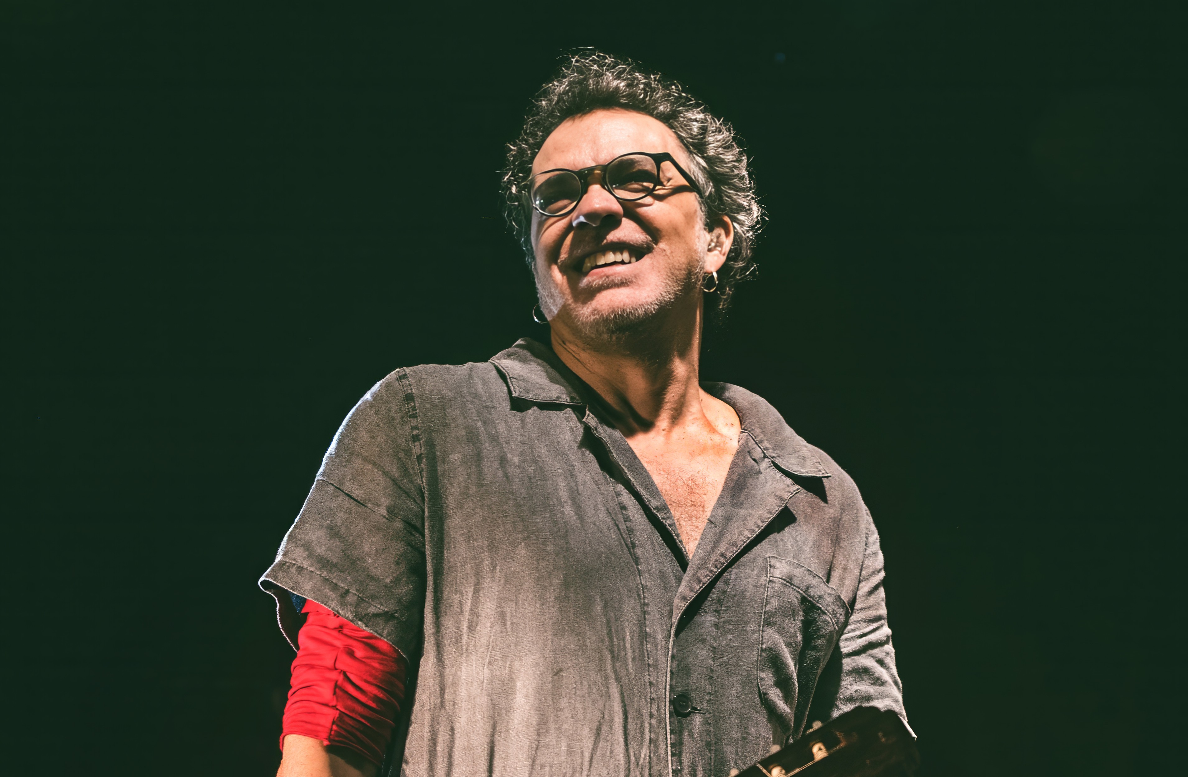 Homem de meia-idade, cabelos grisalhos e óculos, sorrindo e olhando para cima, vestindo camisa cinza sobre blusa vermelha, com brinco na orelha esquerda, em palco escuro