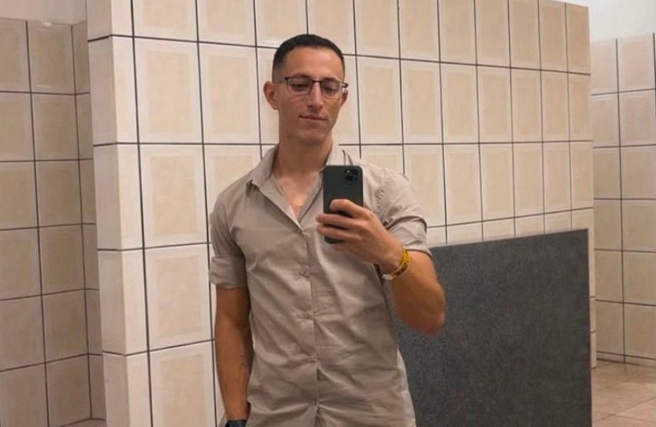 Homem jovem, de óculos e cabelo curto, vestindo camisa bege de manga curta, tira uma selfie no espelho de um banheiro com paredes de azulejos claros e rejunte escuro