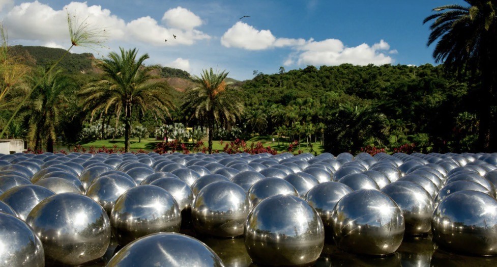 Campo de esferas met&aacute;licas brilhantes refletindo o c&eacute;u azul com nuvens e palmeiras, em um jardim tropical com montanhas verdes ao fundo