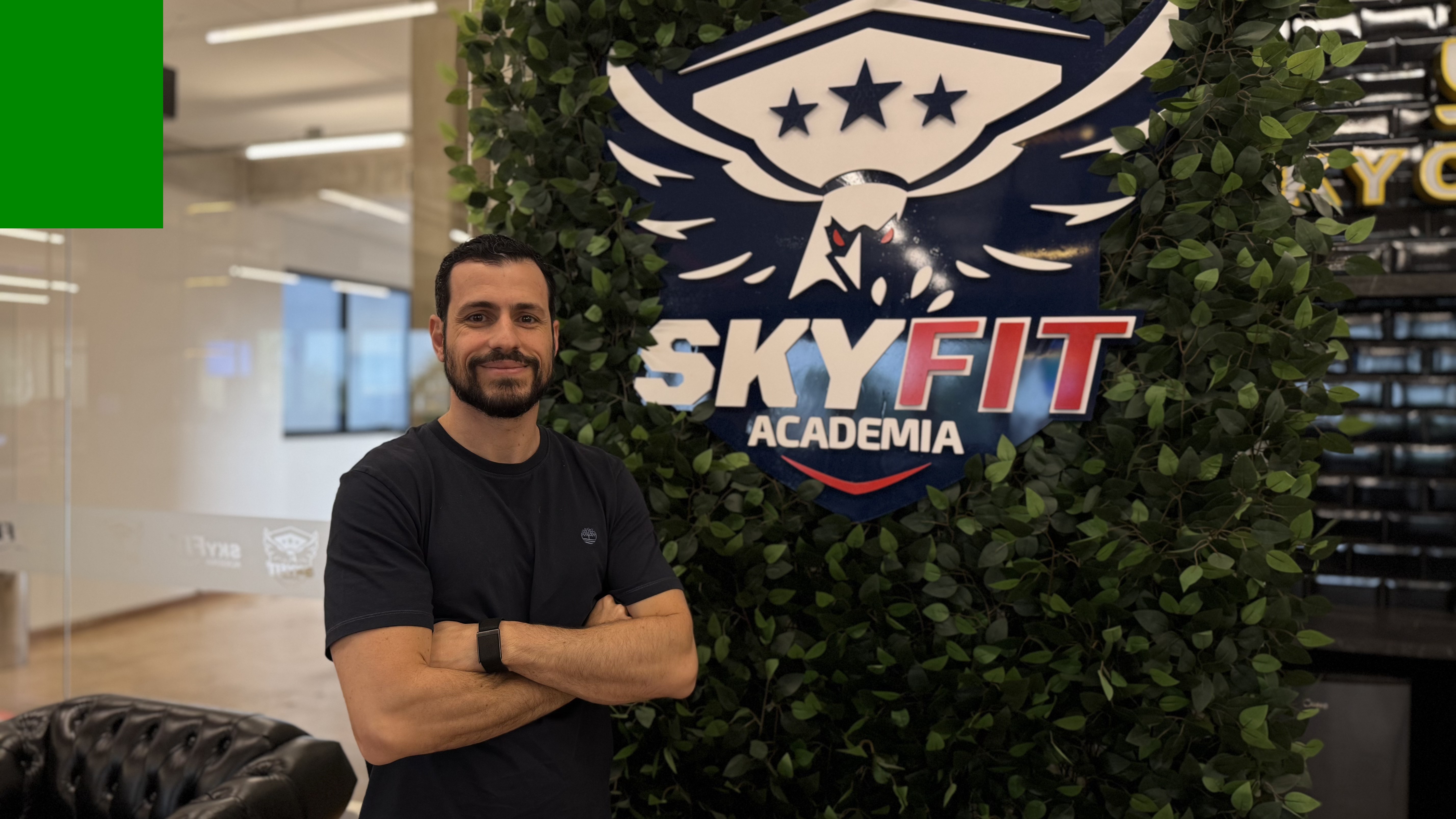 Homem barbudo de camiseta preta e relógio escuro, braços cruzados, sorrindo para a câmera, em frente a um painel verde com o logo da SkyFit Academia