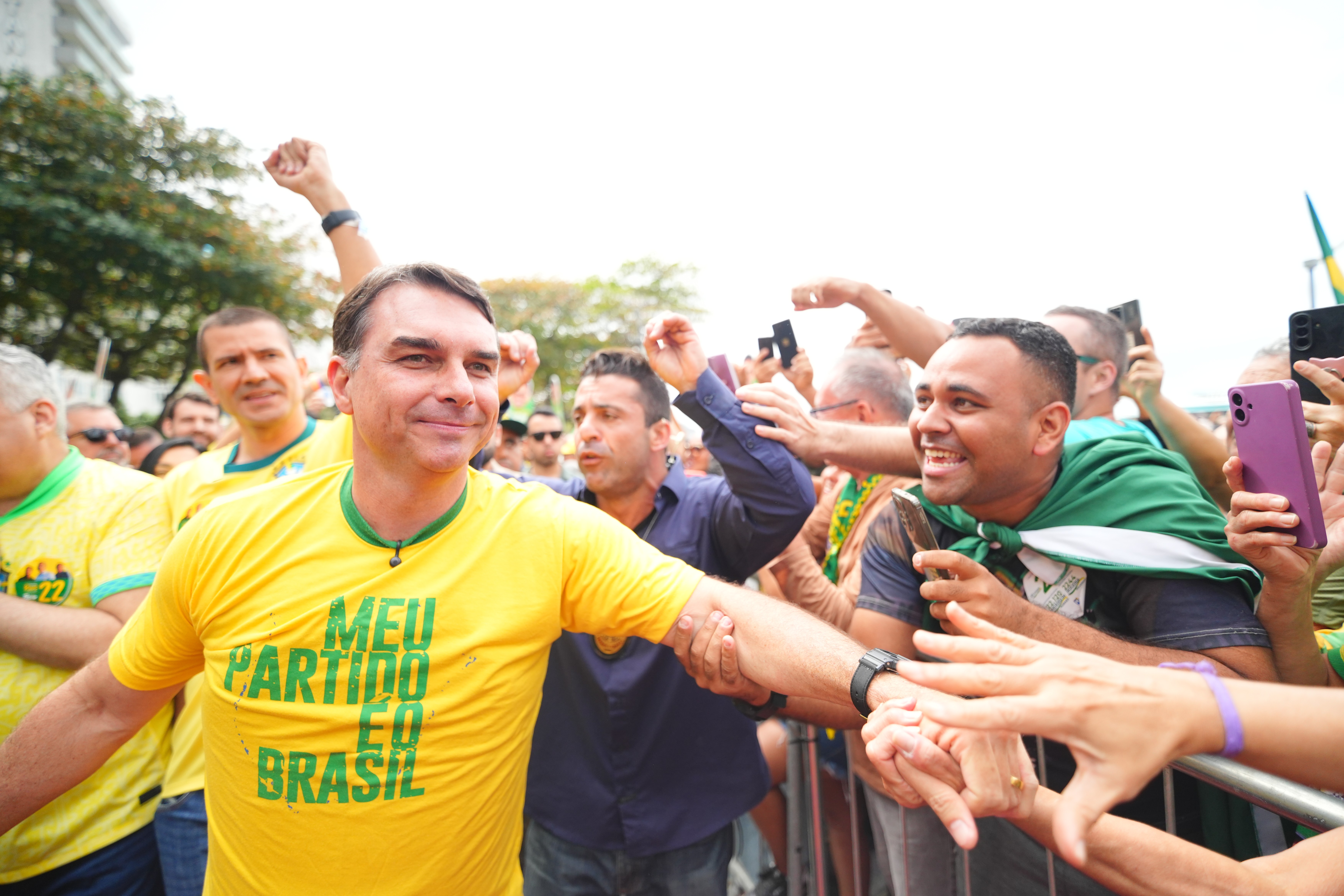 Jair Bolsonaro, sorrindo, cumprimenta apoiadores em evento ao ar livre. Ele veste uma camiseta amarela com a frase MEU PARTIDO É O BRASIL em verde. Ao seu lado, um homem com uma bandeira verde nos ombros sorri, enquanto outros levantam celulares para filmar. O público parece animado, com alguns braços erguidos em sinal de apoio