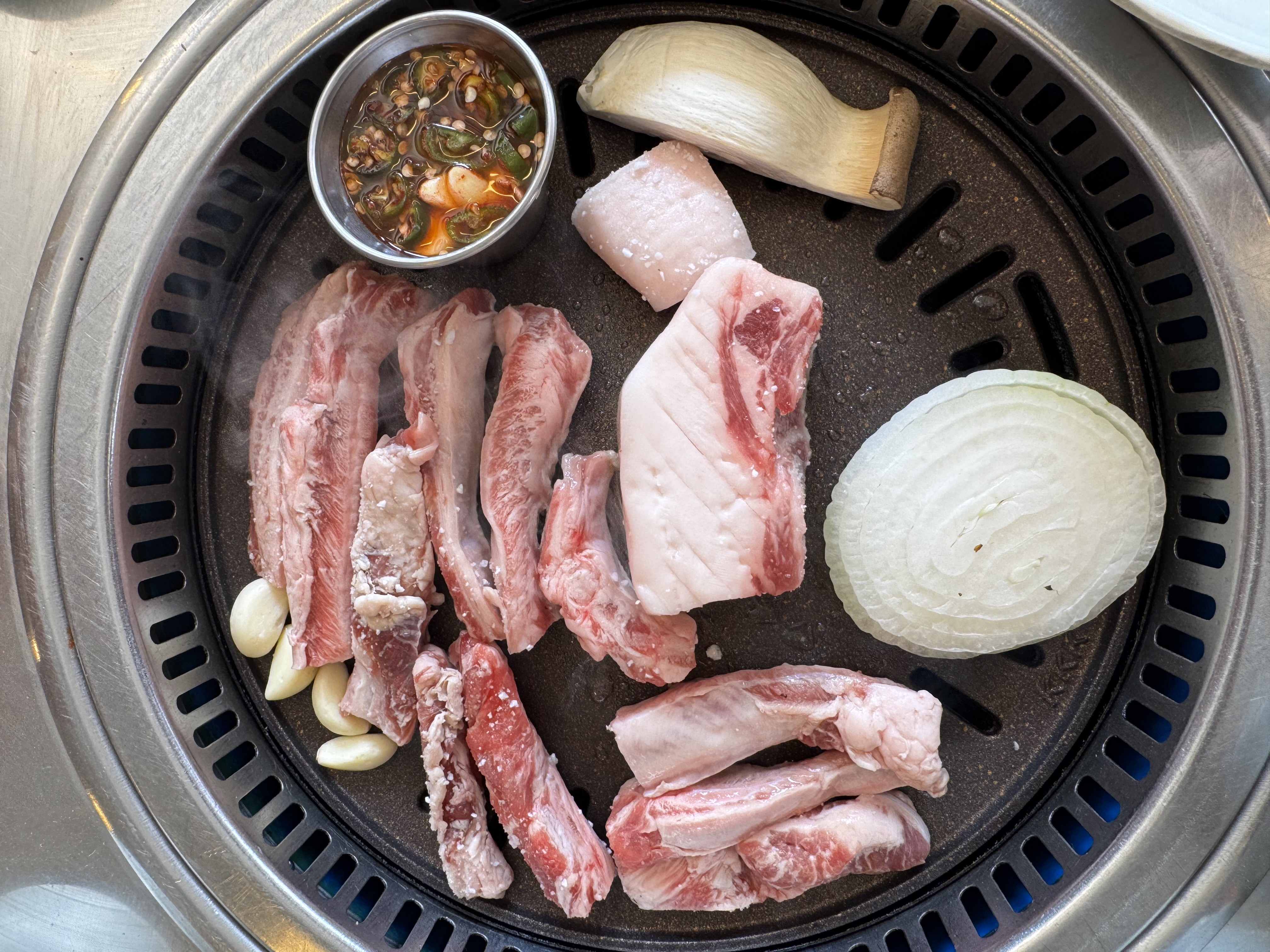 Carnes cruas, costelinhas e peda&ccedil;os de carne de porco, com alho, cebola e um cogumelo, sendo grelhados em uma churrasqueira redonda de metal com uma tigela de molho picante ao lado