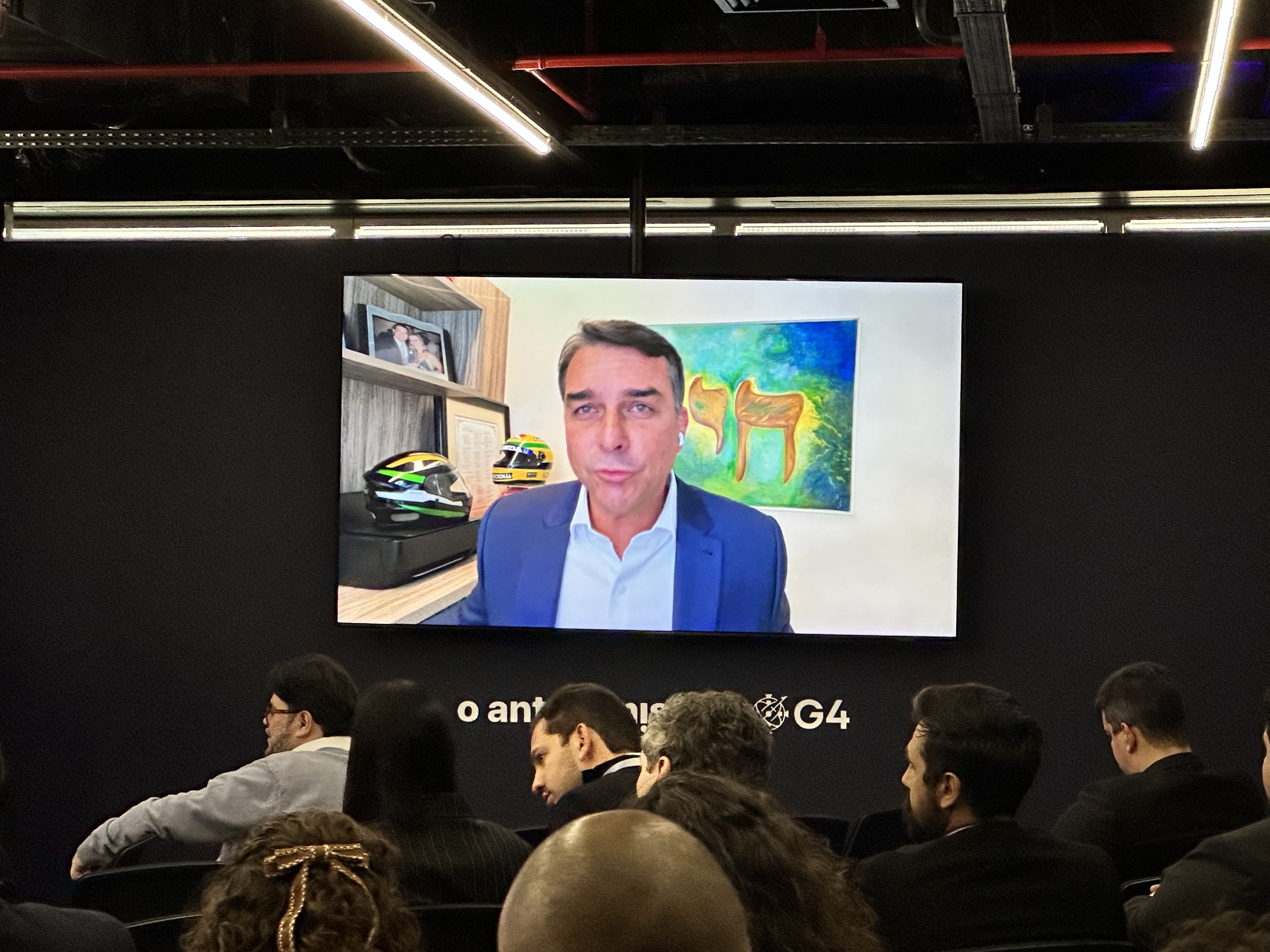 Um homem de meia-idade, com cabelo escuro e terno azul, aparece em uma tela grande, falando para uma plateia em um auditório escuro. Ao fundo da tela, há prateleiras com capacetes e um quadro abstrato. A plateia, vista de costas, observa atentamente a apresentação