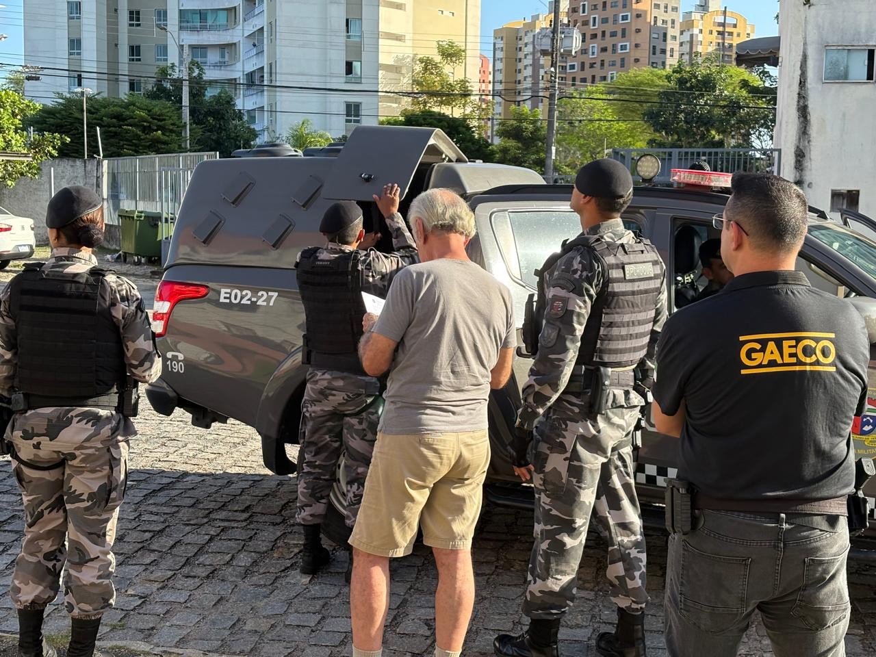 Homem de cabelos grisalhos e camiseta cinza, lendo um documento, cercado por policiais militares fardados e um agente do GAECO, em frente a uma viatura com a caçamba aberta, em rua de paralelepípedos com prédios ao fundo