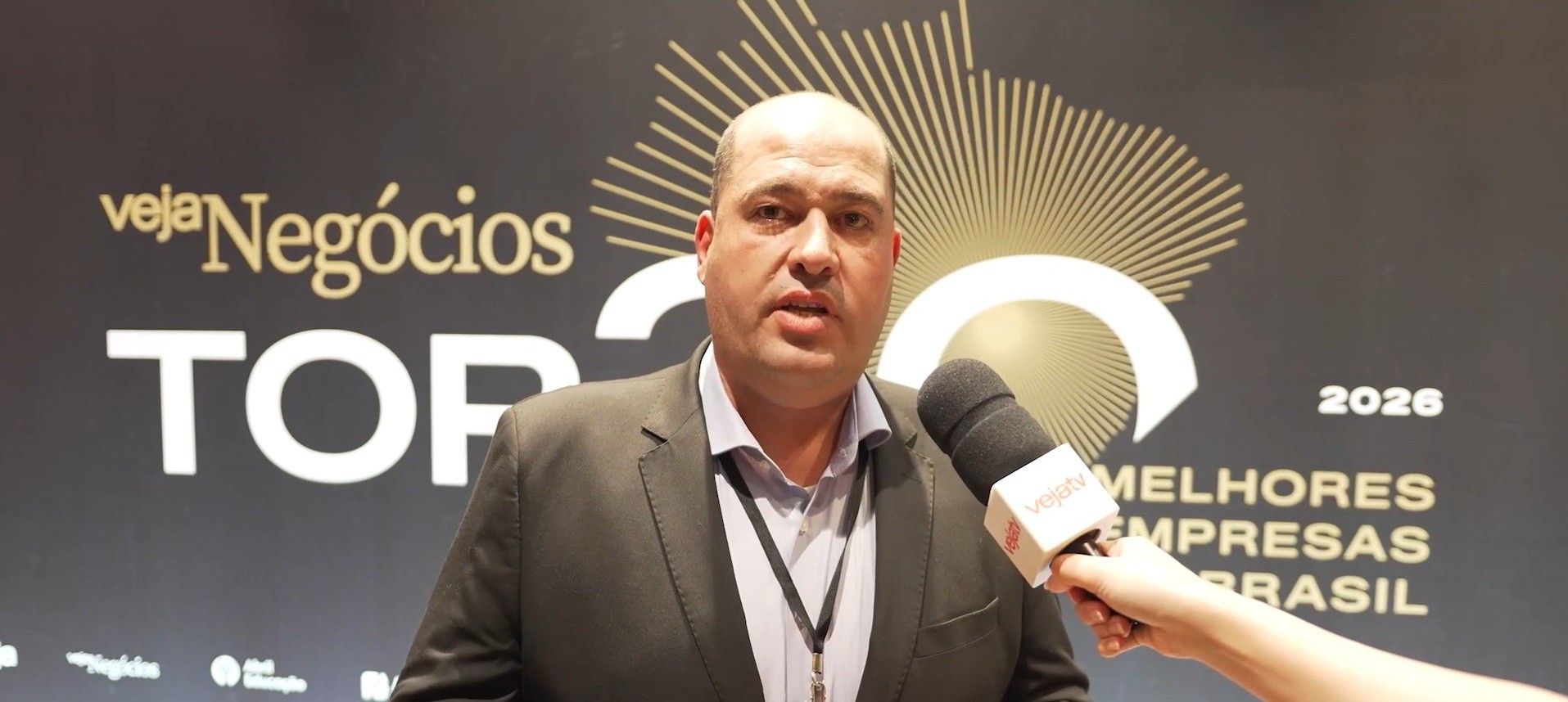 Homem careca de terno cinza e camisa azul claro, com crachá no pescoço, sendo entrevistado por um microfone branco com logo vejatev. Ao fundo, um painel preto com Veja Negócios TOP em letras douradas e brancas, e 2026 MELHORES EMPRESAS BRASIL em dourado