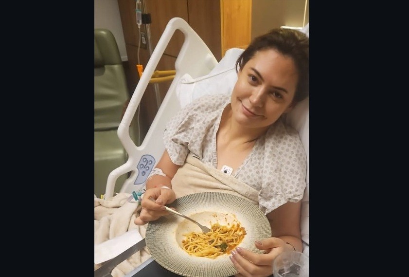 Mulher de cabelos escuros, vestindo camisola hospitalar, sorri levemente enquanto come macarrão com molho vermelho em uma cama de hospital. Ela tem um acesso venoso no braço esquerdo e um monitor cardíaco no peito. Ao fundo, uma poltrona verde e equipamentos médicos