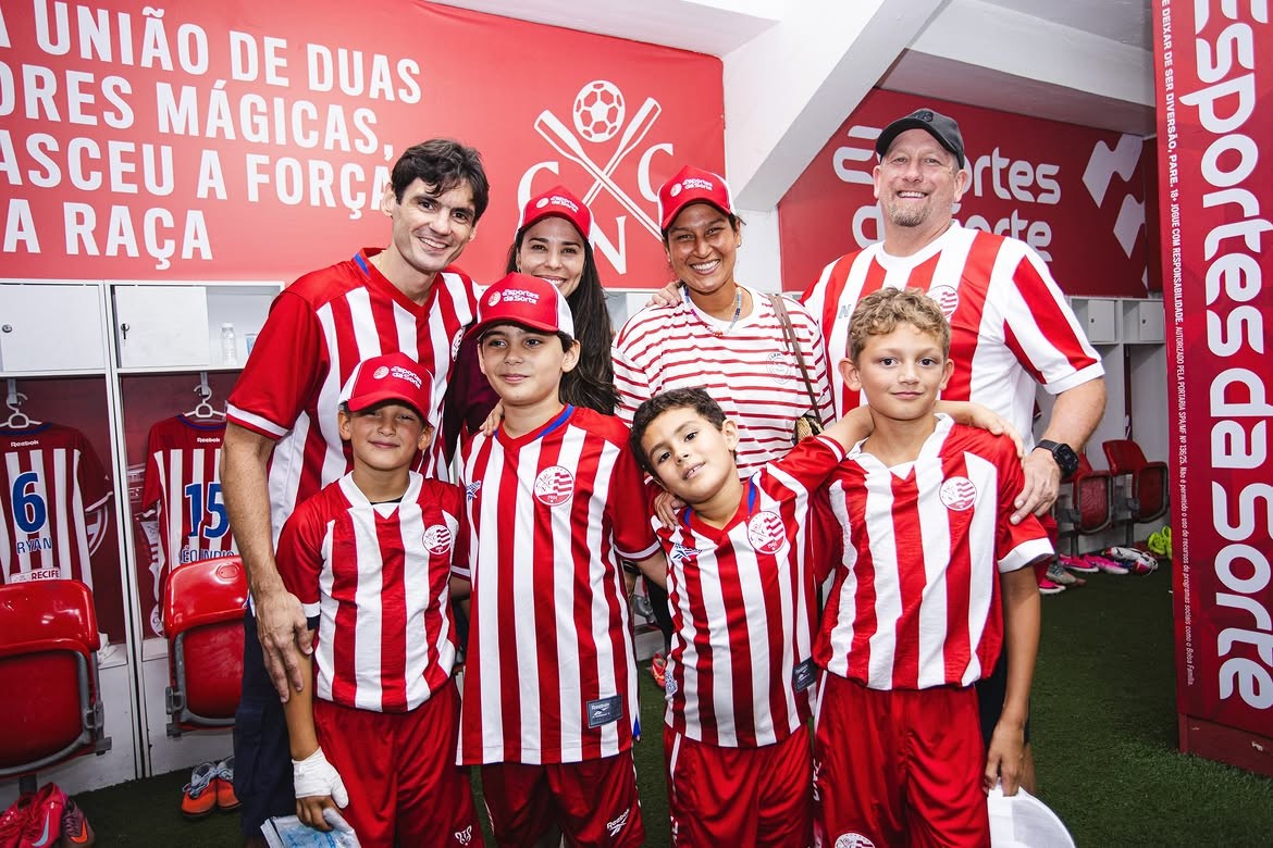 Um grupo de sete pessoas, incluindo quatro crian&ccedil;as e tr&ecirc;s adultos, sorriem para a c&acirc;mera em um vesti&aacute;rio de futebol. Todos vestem camisas listradas vermelhas e brancas, e alguns usam bon&eacute;s vermelhos. Ao fundo, arm&aacute;rios abertos com camisas de futebol penduradas e uma parede vermelha com texto branco e um logotipo de futebol. &Agrave; direita, um banner vertical vermelho com texto branco.