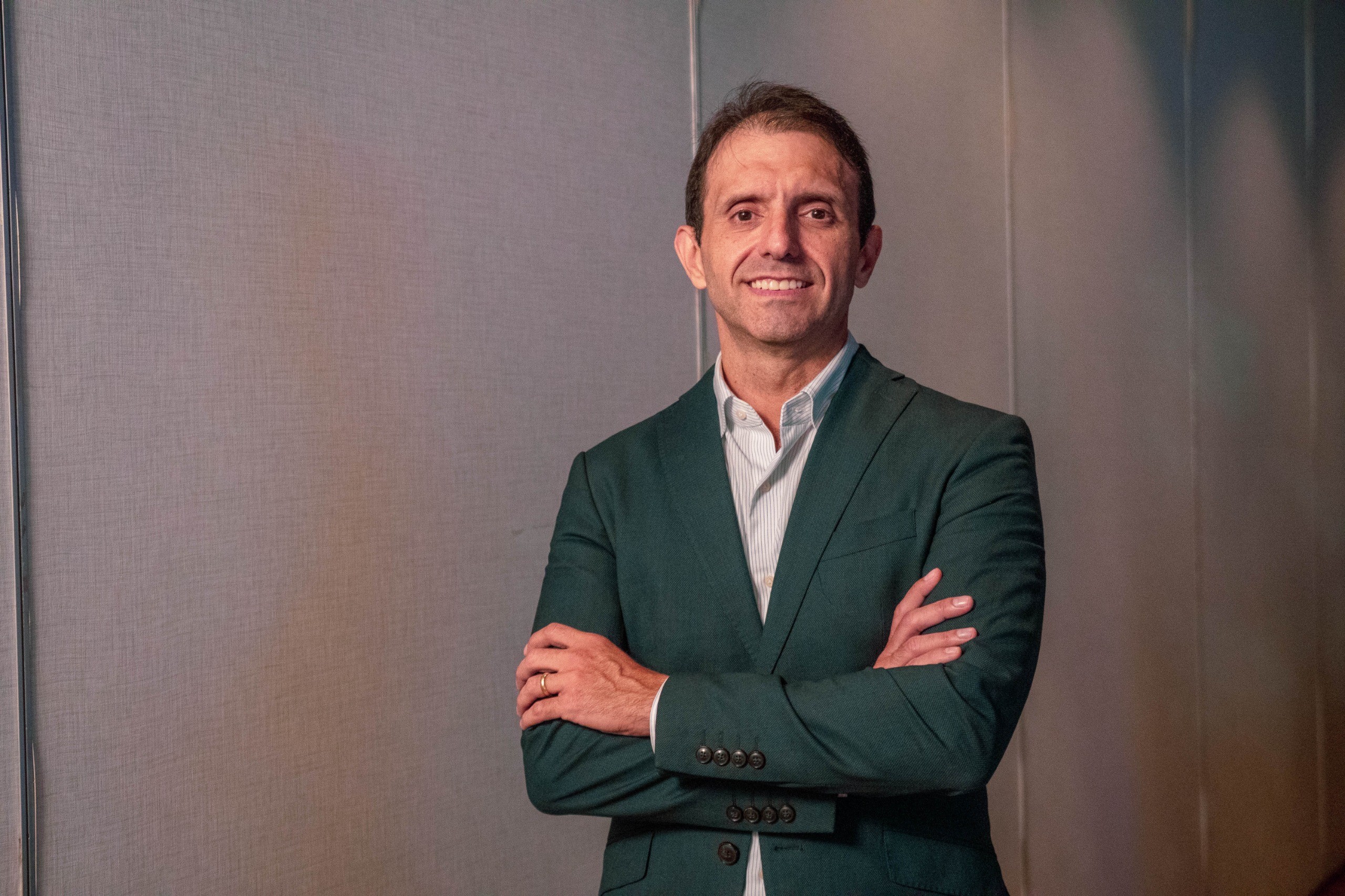 Homem de pele clara, cabelo castanho curto, sorrindo e olhando para frente, veste blazer verde escuro e camisa listrada clara, braços cruzados, em fundo cinza texturizado