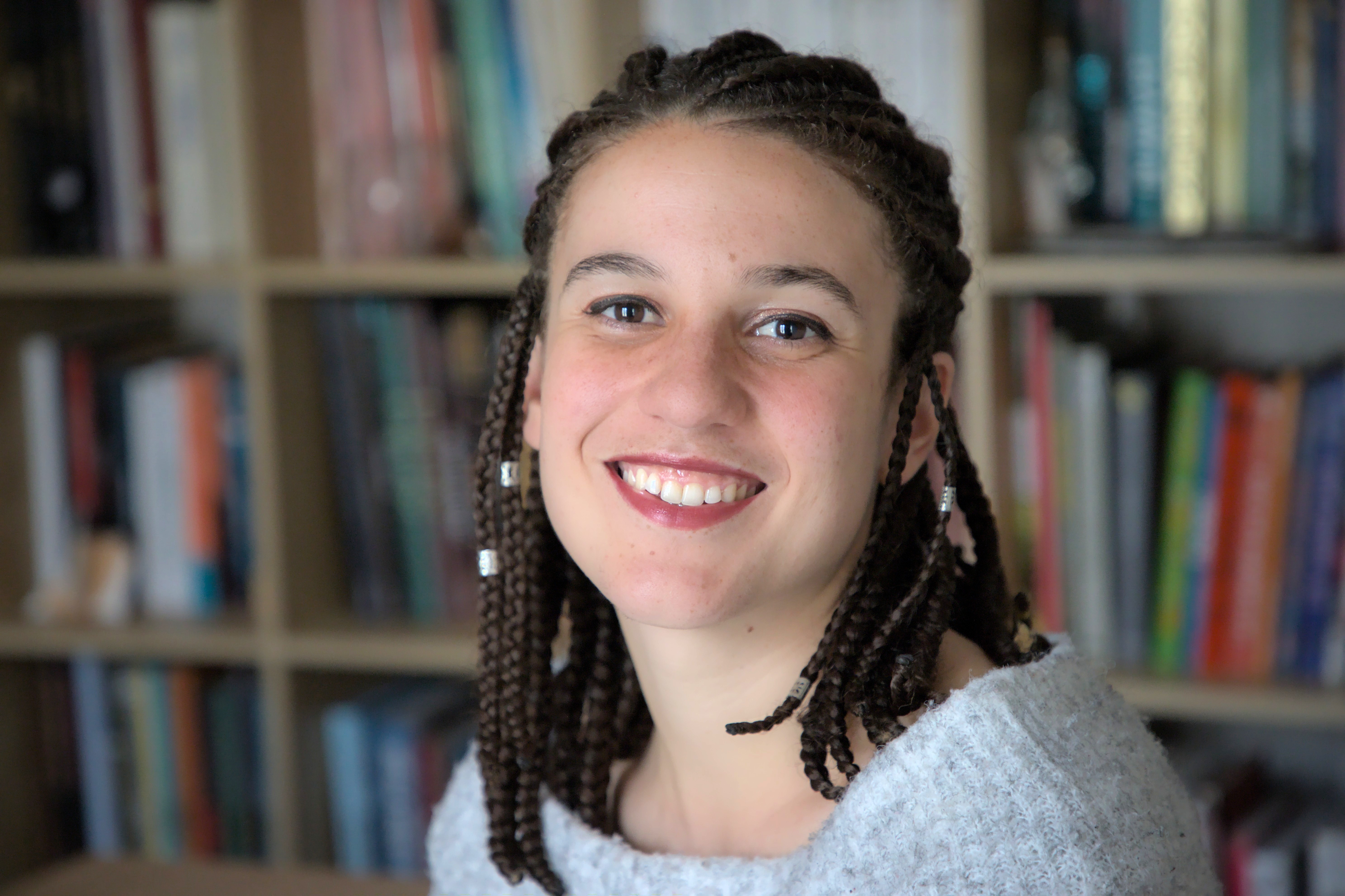 Mulher jovem de pele clara e cabelos castanhos trançados com adornos prateados, sorrindo para a câmera. Veste um suéter cinza e está em frente a uma estante de livros coloridos