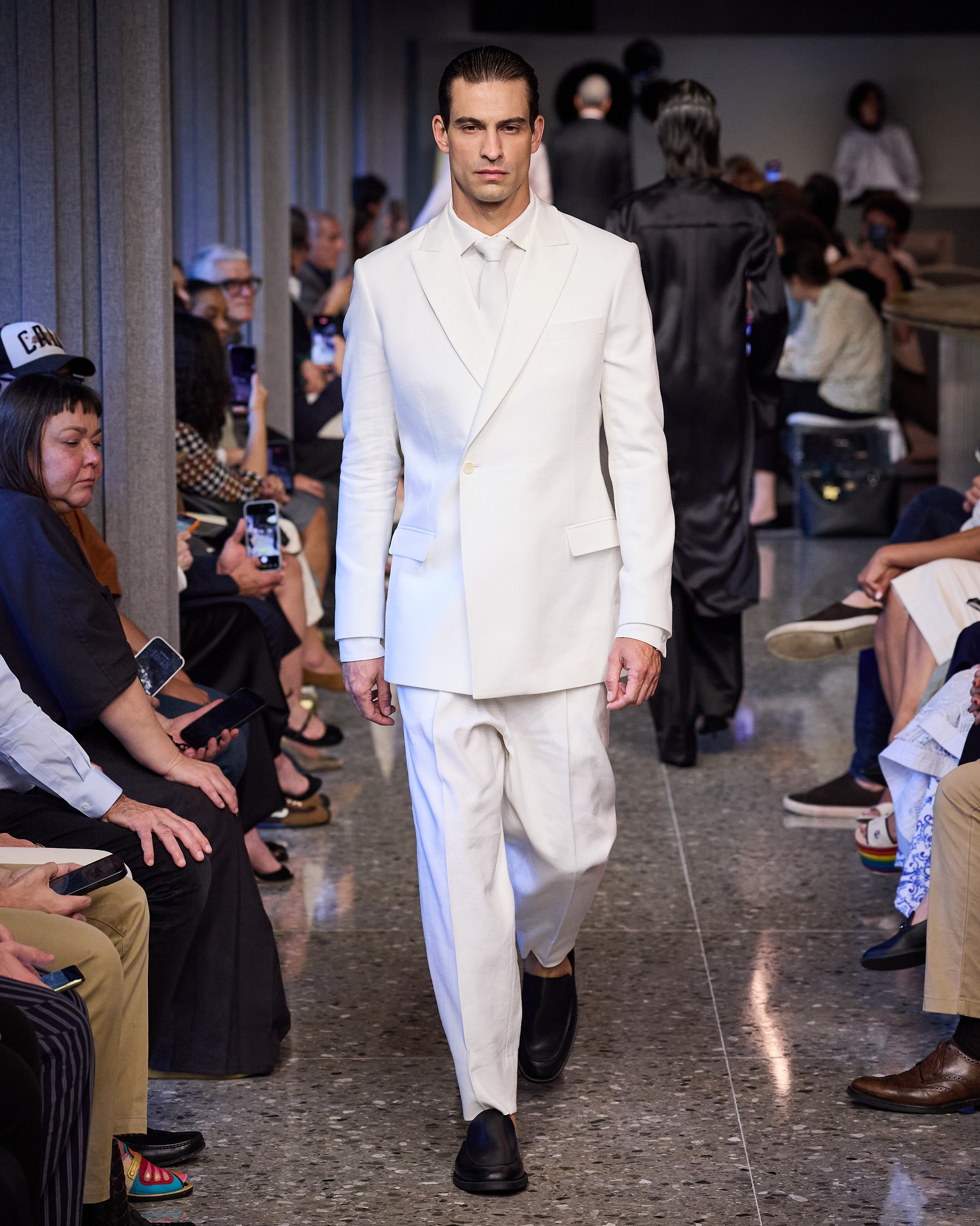 Modelo masculino com cabelo penteado para trás, vestindo um terno branco monocromático com gravata, desfilando em uma passarela. Pessoas sentadas assistem ao desfile, algumas segurando celulares