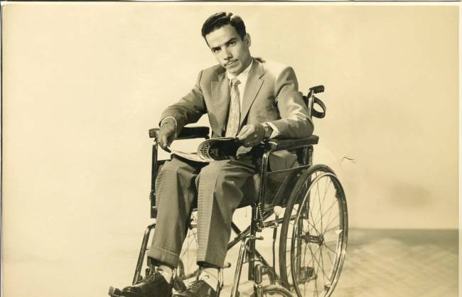 Homem jovem, de terno claro e gravata, sentado em cadeira de rodas, segurando um jornal enrolado. Ele olha diretamente para frente com express&atilde;o s&eacute;ria, em um fundo liso e claro