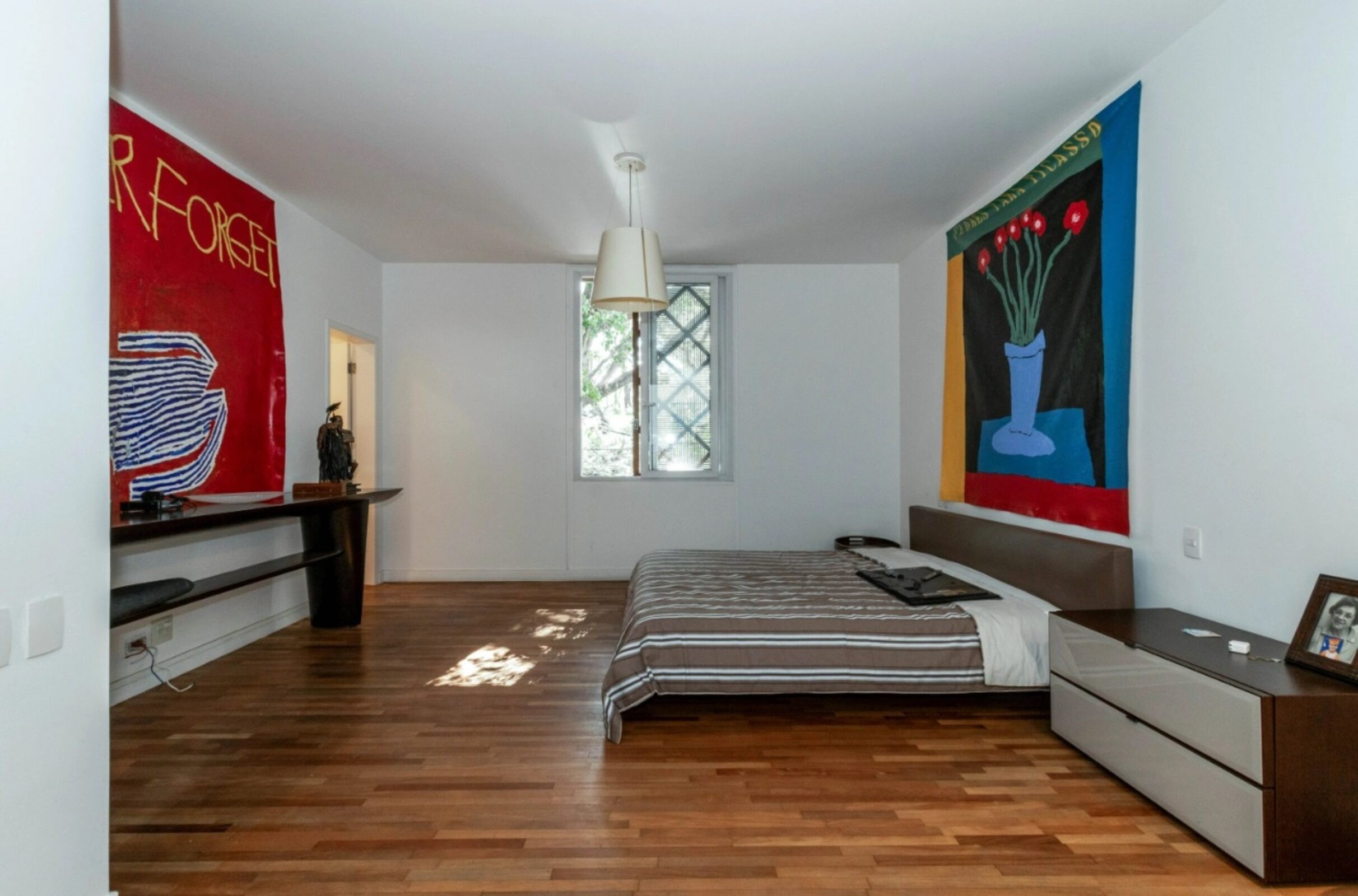 Quarto com paredes brancas, piso de madeira e cama de casal com len&ccedil;ol listrado marrom e branco. H&aacute; uma janela gradeada, um pendente no teto e duas grandes telas coloridas nas paredes: uma vermelha com texto e outra com vaso de flores. Uma mesa de cabeceira escura com gavetas claras e um porta-retrato est&aacute; &agrave; direita da cama