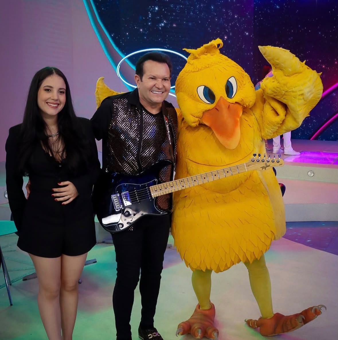 Uma mulher, um homem e um personagem de pato amarelo posam sorrindo. A mulher, à esquerda, veste um macacão preto. O homem, ao centro, usa colete brilhante e segura uma guitarra preta. O pato, à direita, tem bico laranja e olhos azuis, com uma das asas levantada. O fundo é um cenário de palco com luzes coloridas e estrelas