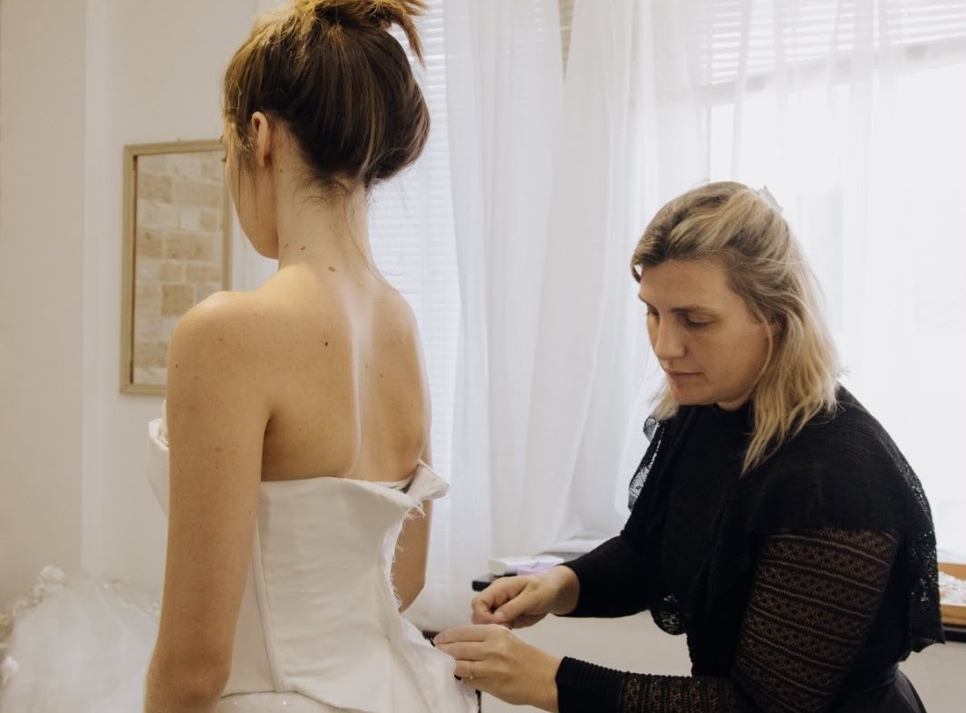 Uma mulher de costas, com cabelo preso, experimenta um vestido de noiva branco tomara que caia, enquanto uma costureira loira, de blusa preta, ajusta a peça com as mãos. O cenário é um ateliê claro com uma janela e cortinas brancas.