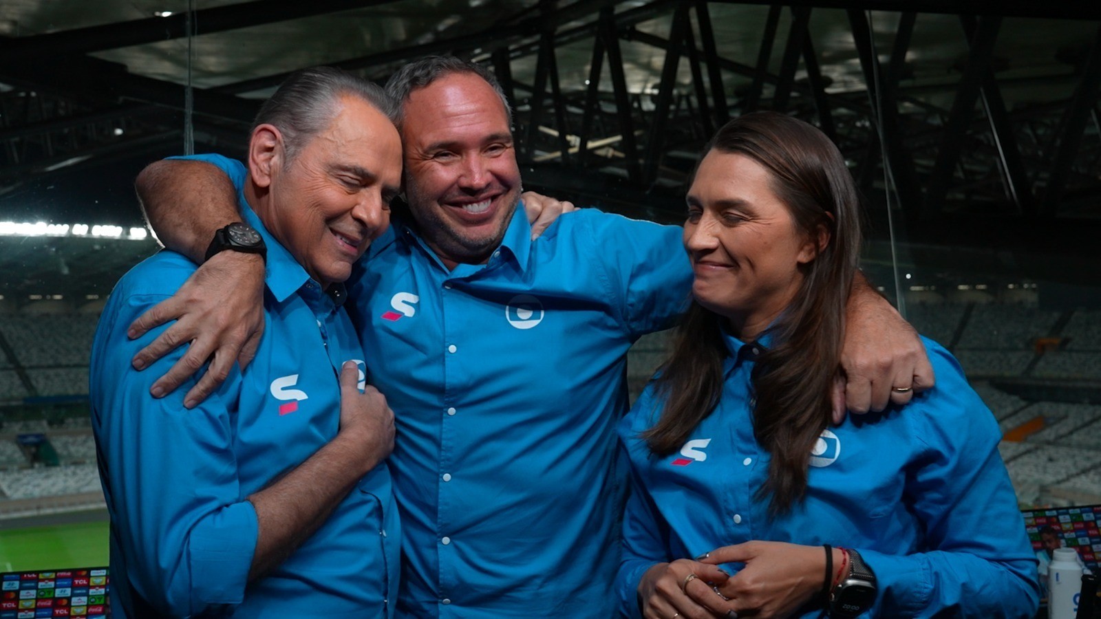 Três pessoas sorridentes, vestindo camisas sociais azuis com logotipos, abraçadas em um estádio com arquibancadas ao fundo