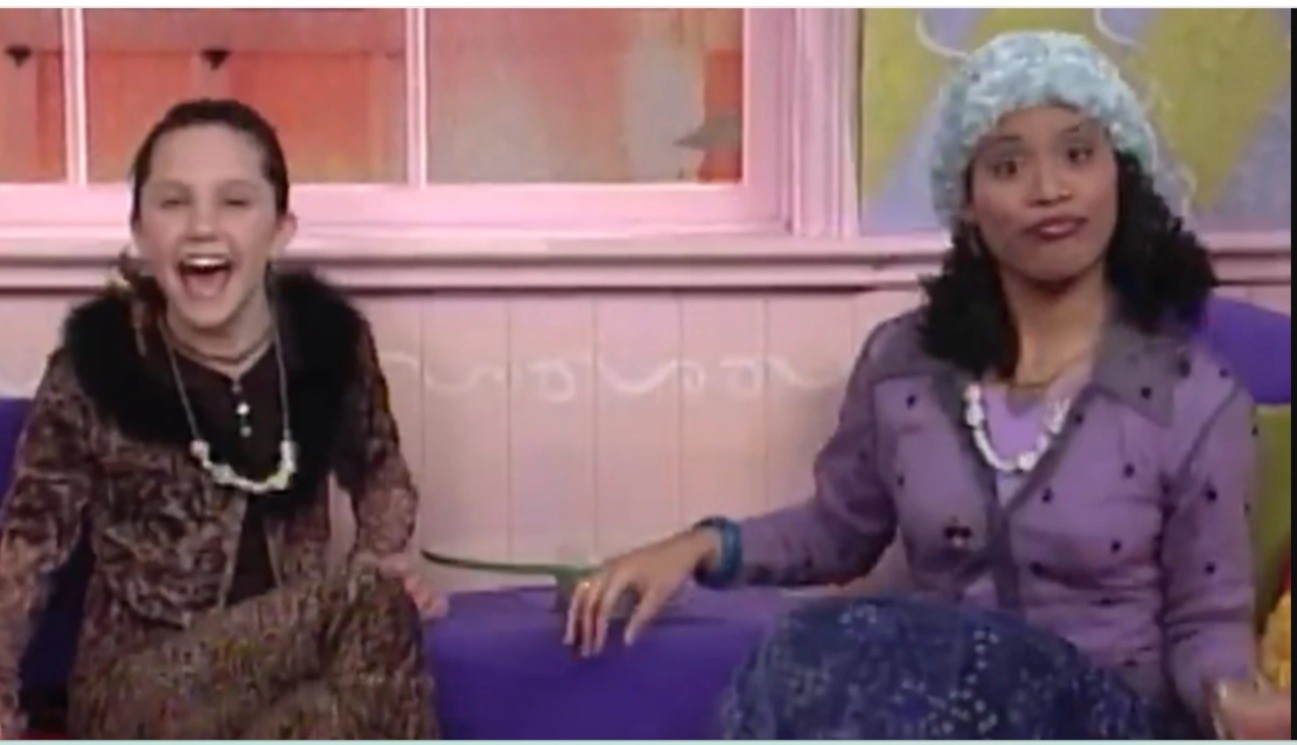 Duas mulheres sentadas em um sofá roxo. À esquerda, uma jovem de cabelo escuro preso, blusa marrom com estampa de onça e gola de pelo, gargalha com a boca aberta. À direita, uma mulher de pele escura, cabelo escuro, usa um gorro azul claro e um casaco lilás com bolinhas escuras, olhando para frente com expressão neutra