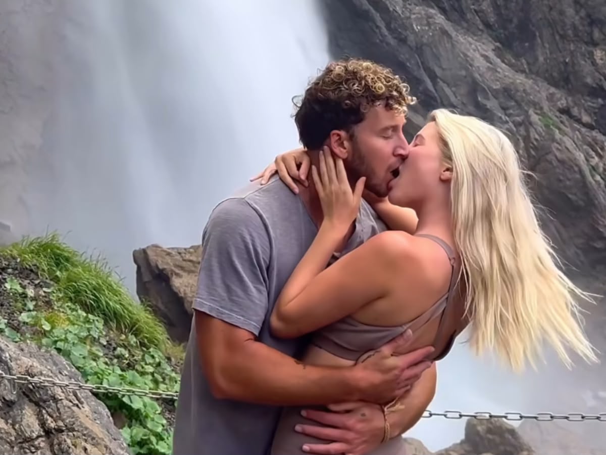 Um casal se beijando apaixonadamente em frente a uma cachoeira. O homem, de cabelo cacheado e barba, veste camiseta cinza. A mulher, loira de cabelo comprido, usa top e calça marrons. Ambos se abraçam, com as mãos dela no pescoço dele e as mãos dele na cintura dela. Ao fundo, rochas e vegetação verde.