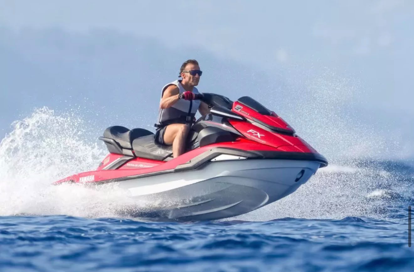Homem de óculos escuros e colete salva-vidas cinza e branco pilotando um jet ski vermelho e branco sobre a água azul, levantando respingos