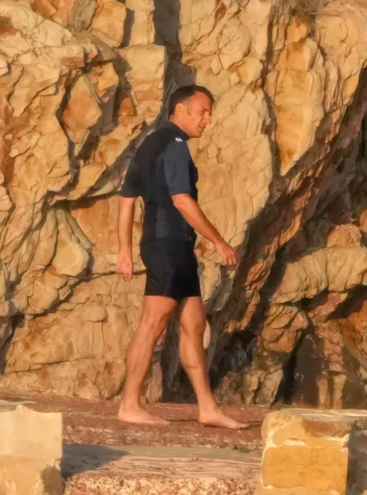 Homem de pele clara, cabelo escuro curto, vestindo camisa polo azul-marinho e bermuda escura, caminhando descalço sobre pedras claras, com uma parede rochosa de tons terrosos ao fundo