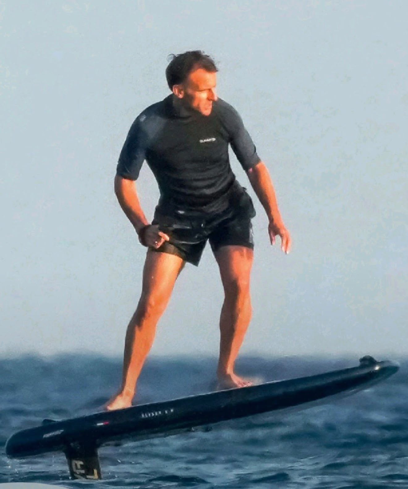 Emmanuel Macron, presidente da França, em pé sobre uma prancha de surf preta com hidrofólio, flutuando sobre a água azul do mar. Ele veste uma camisa de manga curta e shorts pretos, com expressão concentrada, olhando para a direita. Seus braços estão levemente flexionados, equilibrando-se. O céu claro e azulado forma o fundo.