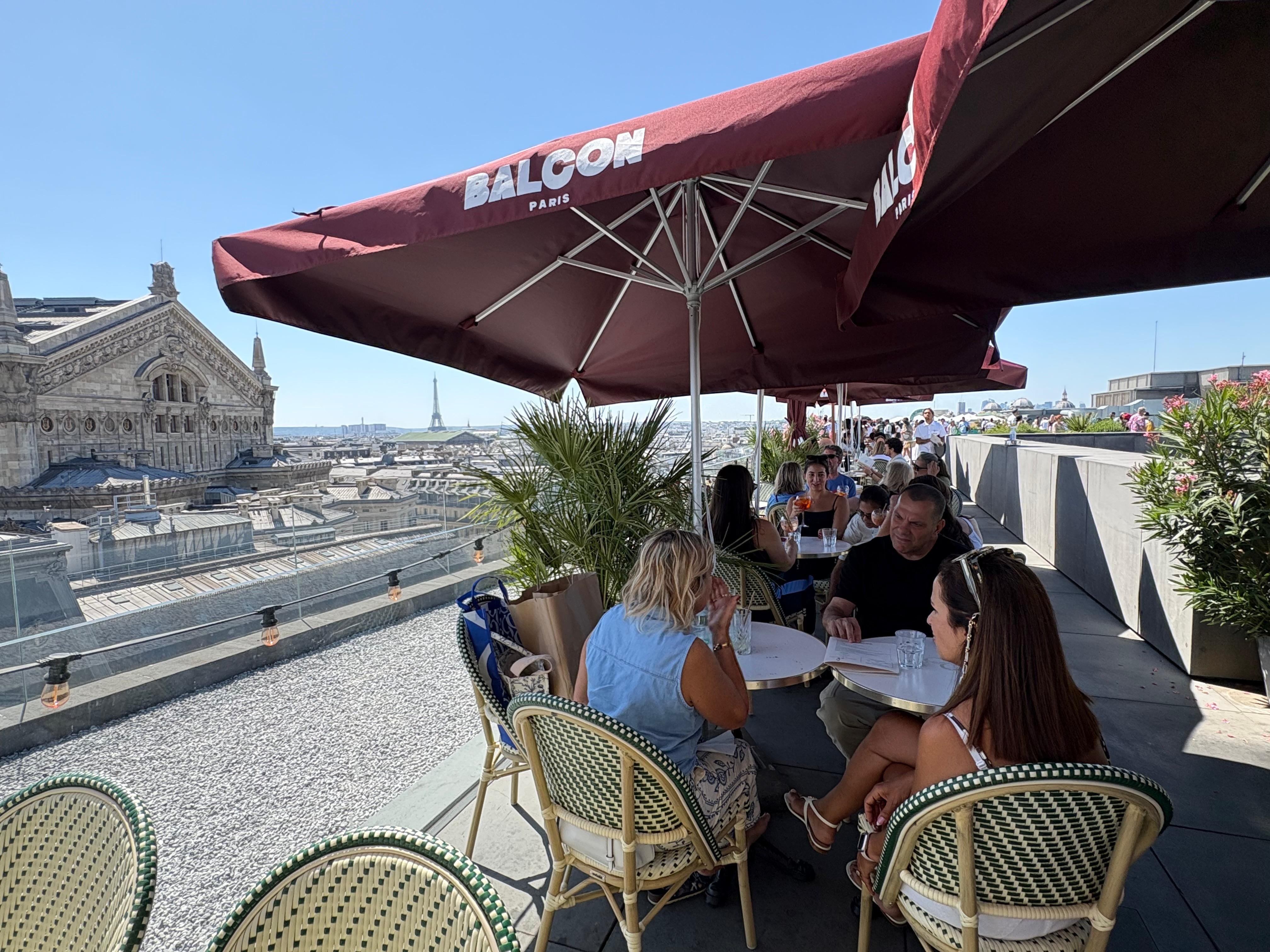 Pessoas sentadas em mesas de um restaurante ao ar livre no terraço de um prédio em Paris, com a Torre Eiffel ao fundo. Guarda-sóis grandes e vermelhos cobrem as mesas, e o céu está azul e claro.