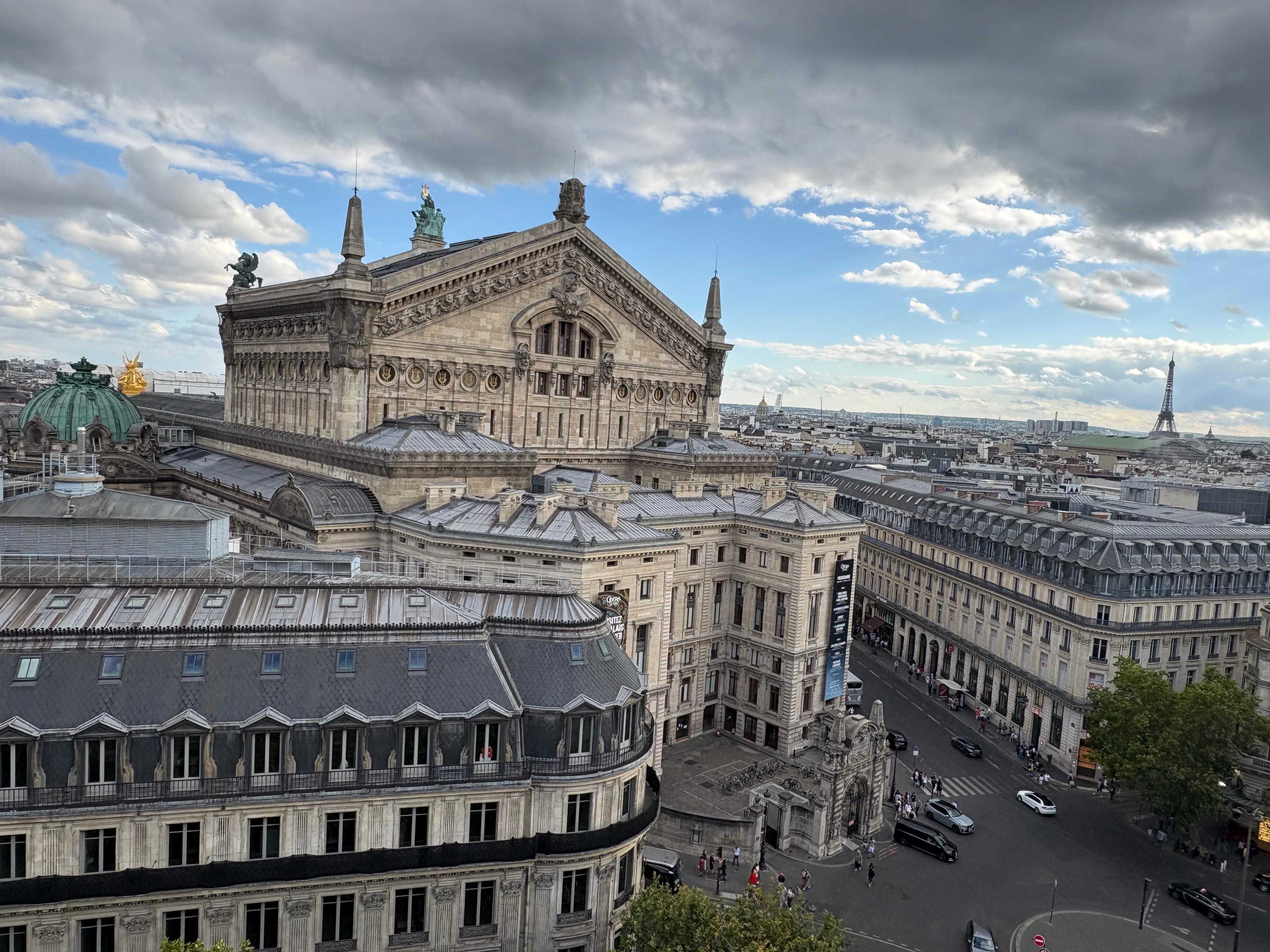 Vista aérea do Palais Garnier, um edifício histórico de Paris com telhados cinzas e cúpulas verdes, sob um céu parcialmente nublado. Ao fundo, a Torre Eiffel se destaca na paisagem urbana.