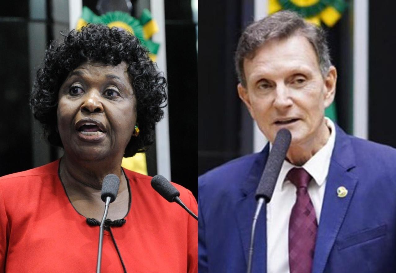 Duas pessoas em primeiro plano, uma mulher negra de cabelo cacheado e blusa vermelha falando ao microfone, e um homem branco de terno azul e gravata vinho também falando ao microfone