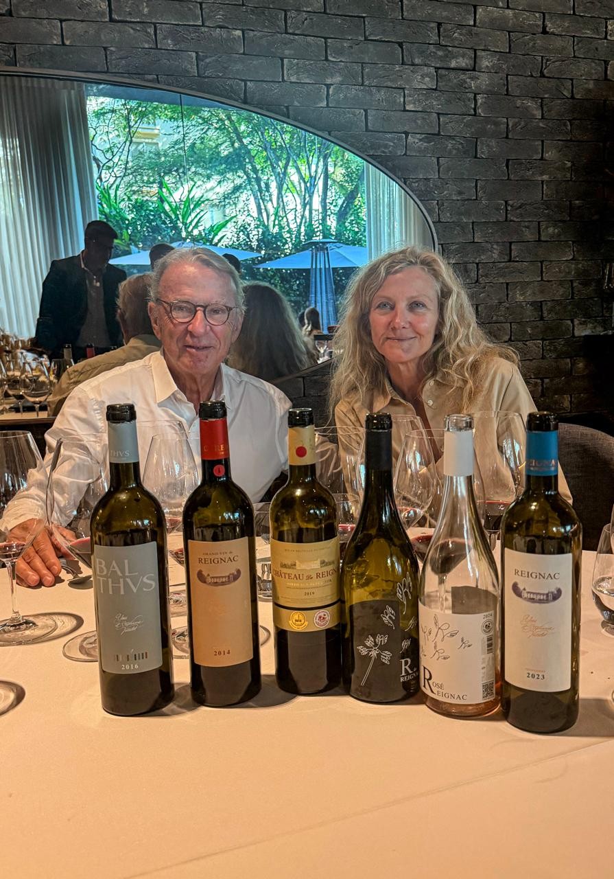 Um homem e uma mulher sorriem para a câmera, sentados à mesa de um restaurante com seis garrafas de vinho e taças. O homem usa óculos e camisa branca, a mulher tem cabelos loiros e veste camisa bege. Ao fundo, um espelho reflete árvores e um guarda-sol azul
