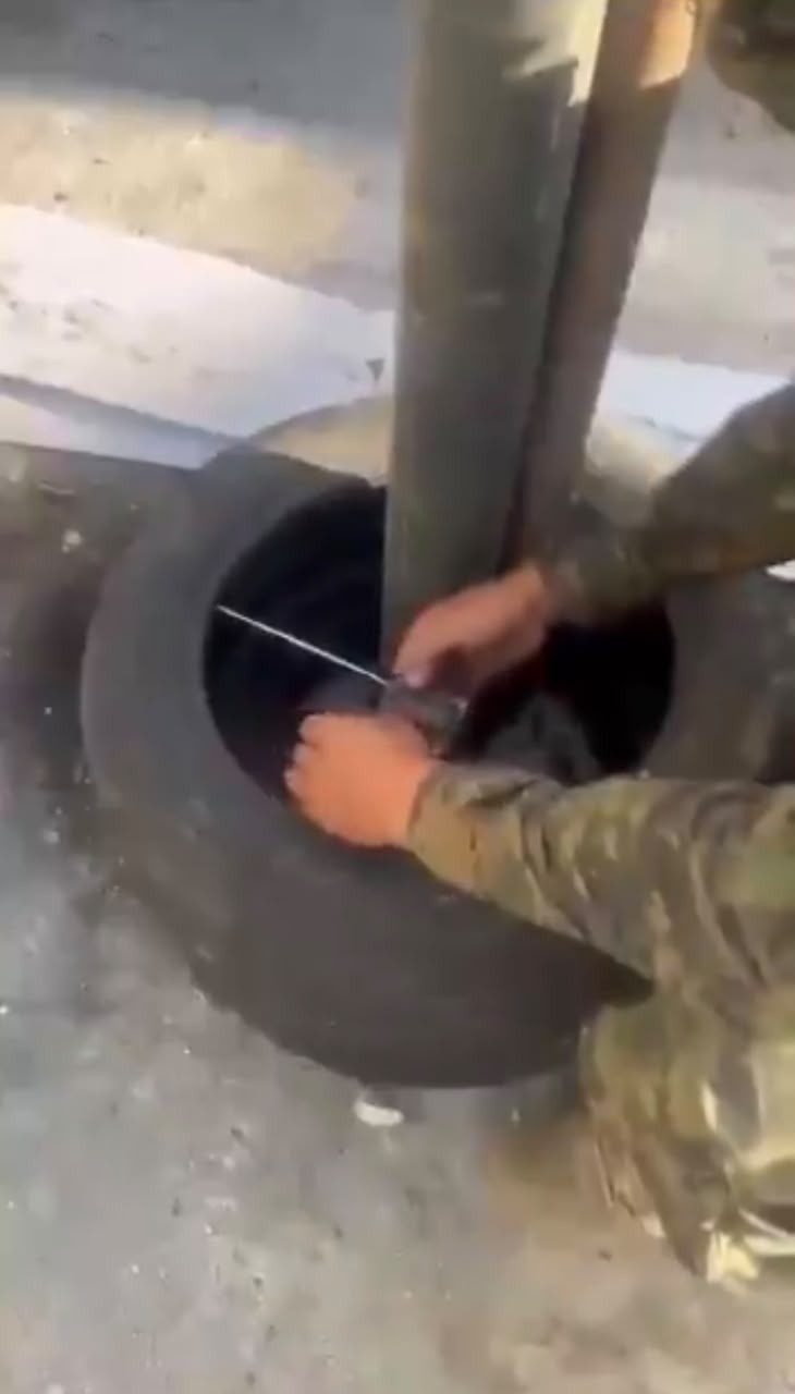Duas mãos vestindo uniforme camuflado inserem um objeto metálico fino em um pneu preto, que está em pé no chão de concreto, ao lado de um poste cinza