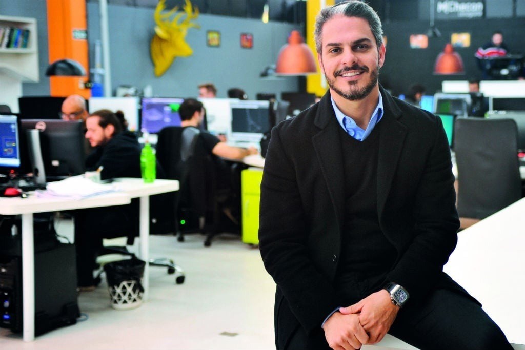 Homem branco de barba, cabelo grisalho e sorriso, vestindo blazer preto e camisa azul, sentado em uma mesa de escritório com as mãos cruzadas. Ao fundo, colegas trabalham em computadores, com decoração moderna e luminárias pendentes