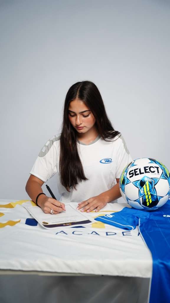 Jovem mulher de cabelos longos e escuros, vestindo camiseta esportiva branca, assina um documento sobre uma toalha de mesa branca com detalhes amarelos e azuis. Ao lado, uma bola de futebol SELECT e uma camisa azul dobrada. Ela est&aacute; concentrada na assinatura