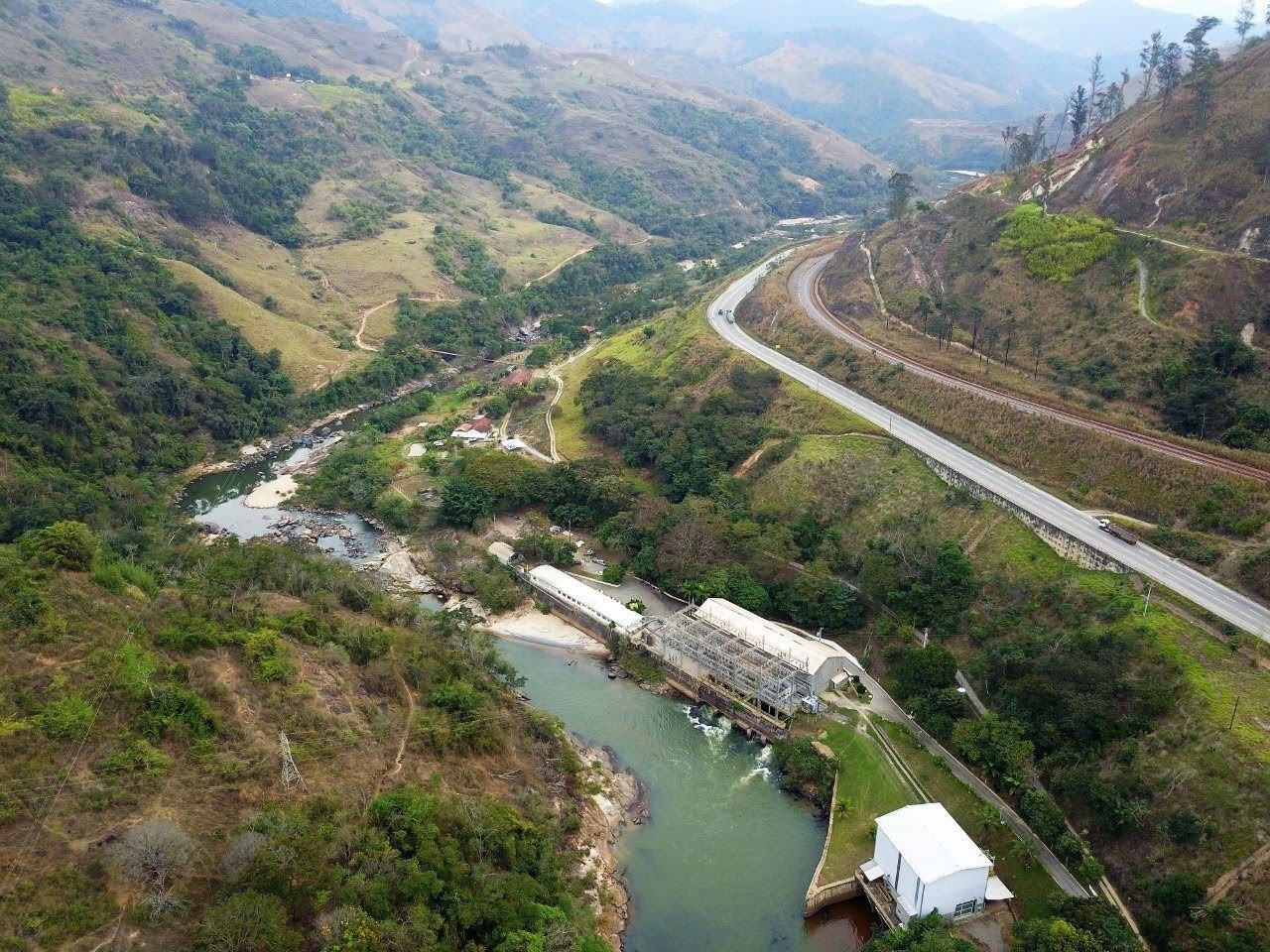 Vista aérea de uma usina hidrelétrica com telhado branco e estrutura metálica cinza, à beira de um rio verde-escuro. Ao lado, uma estrada sinuosa e uma linha férrea cortam a paisagem montanhosa coberta por vegetação densa. O céu está nublado, e o ambiente transmite tranquilidade.