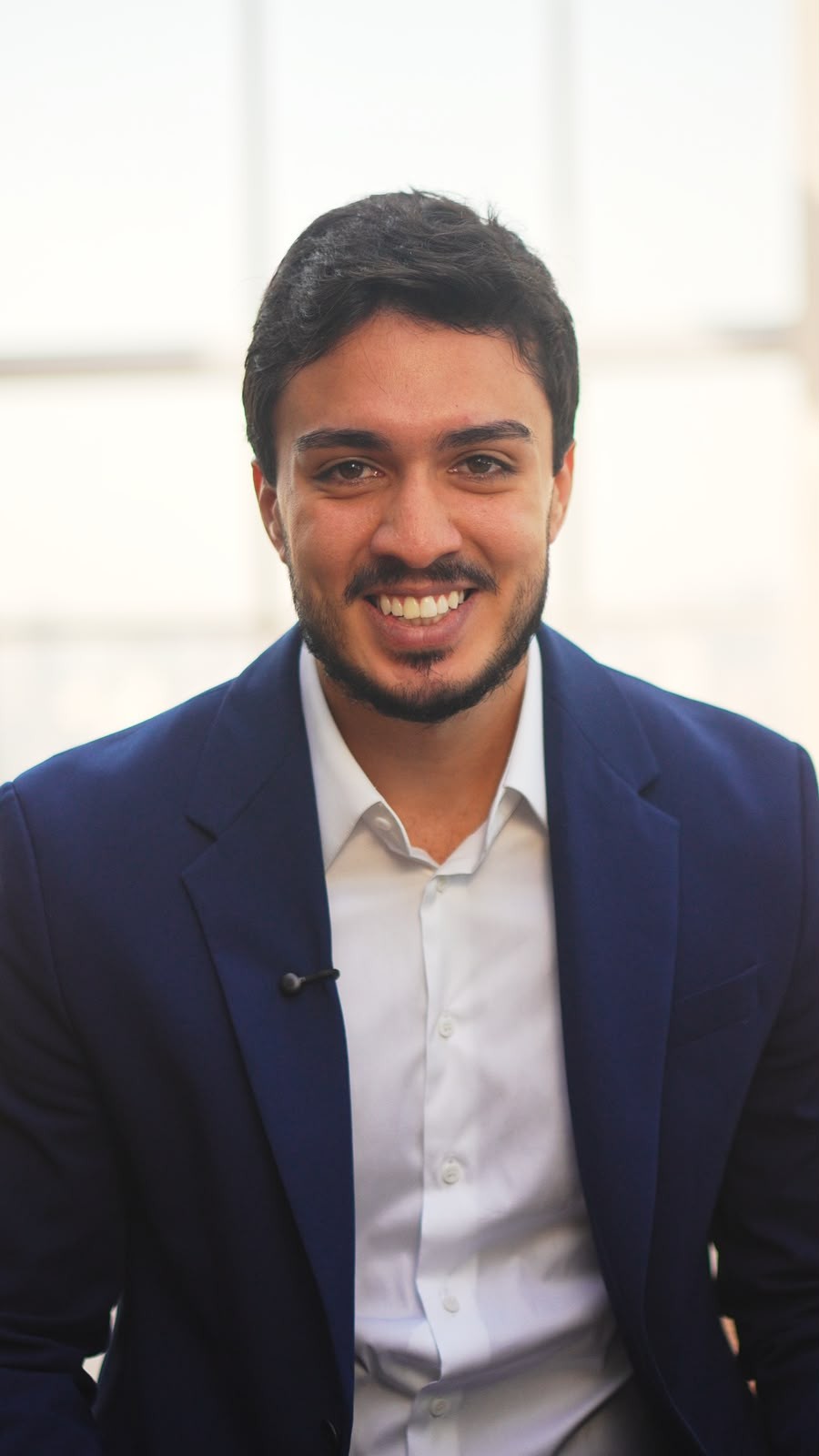 Homem jovem, pele morena, cabelo e barba escura, sorrindo para a c&acirc;mera. Veste blazer azul-marinho e camisa social branca, com um microfone de lapela preso ao blazer. Fundo claro e desfocado