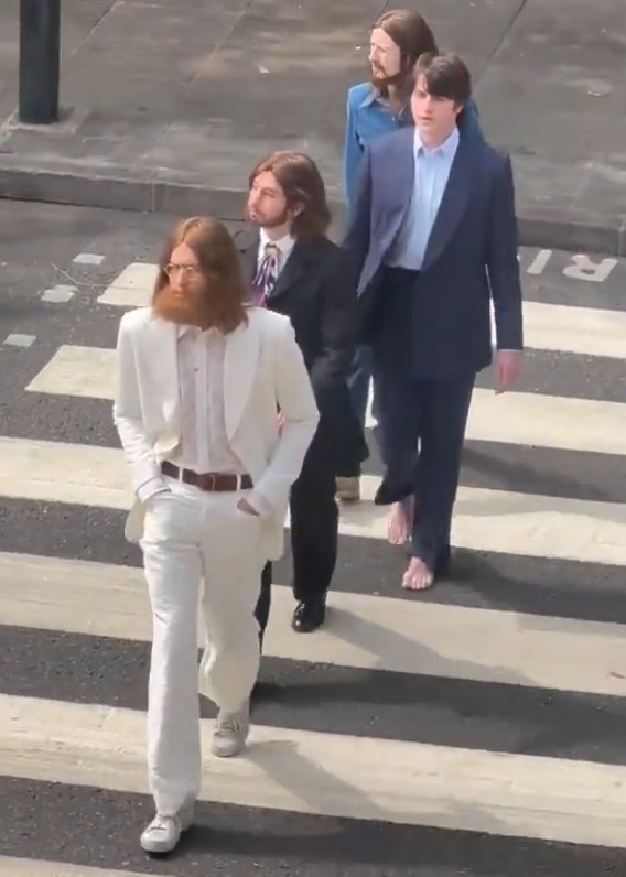 Quatro homens, caracterizados como os Beatles, atravessam uma faixa de pedestres. O primeiro, de barba e óculos, veste terno branco. O segundo, de terno preto e gravata roxa. O terceiro, de terno azul e camisa clara, está descalço. O último, de camisa jeans, está atrás. A cena remete à capa do álbum Abbey Road