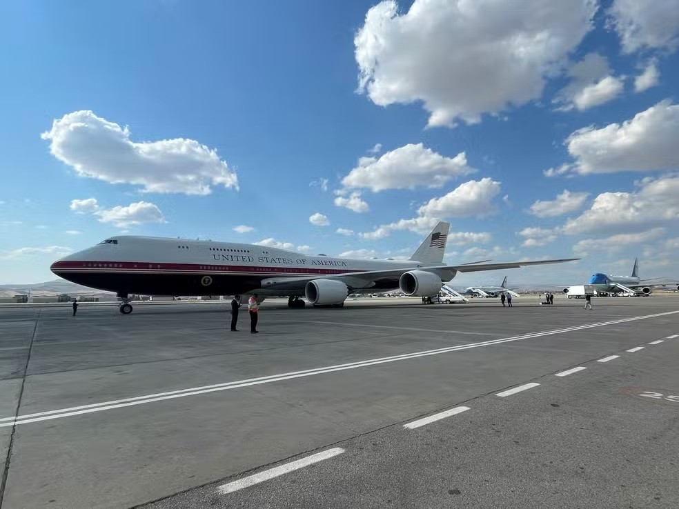 Avião presidencial Air Force One, branco com faixa azul e vermelha, estacionado em pista de aeroporto sob céu azul com nuvens esparsas. Pessoas pequenas estão próximas à aeronave, e outros aviões menores aparecem ao fundo.