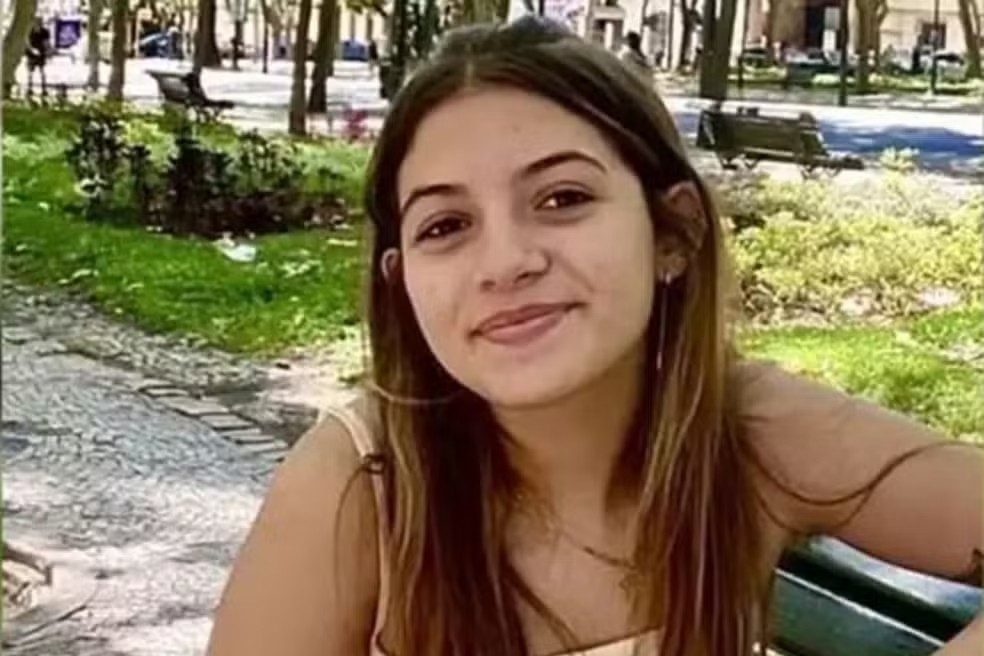 Jovem mulher de cabelos castanhos longos, sorrindo levemente, sentada em um banco de parque com blusa bege de alças finas. Ao fundo, árvores e gramado verde