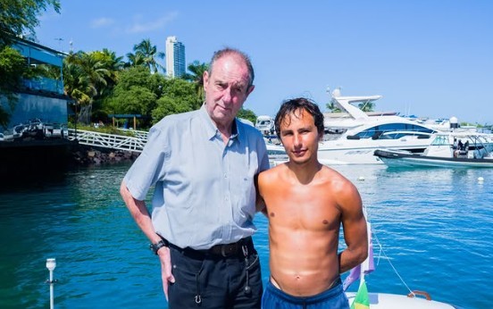 Dois homens, um mais velho de camisa azul clara e cal&ccedil;a preta, e um jovem sem camisa com sunga azul, posam em um p&iacute;er. Ao fundo, barcos e iates na &aacute;gua, com vegeta&ccedil;&atilde;o e um pr&eacute;dio alto sob c&eacute;u azul