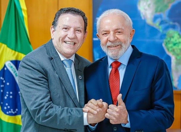 Dois homens sorridentes, um de terno cinza e gravata azul, outro de terno azul e gravata vermelha, apertam as mãos. Ao fundo, a bandeira do Brasil e um mapa-múndi