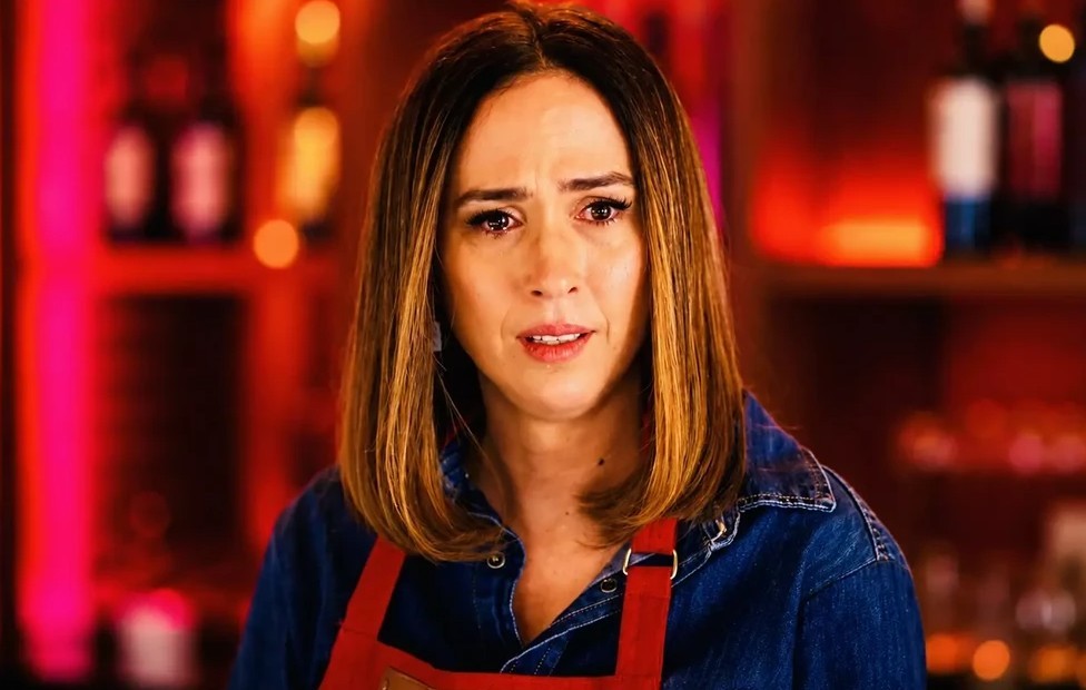 Mulher de cabelos castanhos claros, com expressão de choro e olhos marejados, vestindo camisa jeans azul e avental vermelho, em ambiente com luzes vermelhas e garrafas desfocadas ao fundo