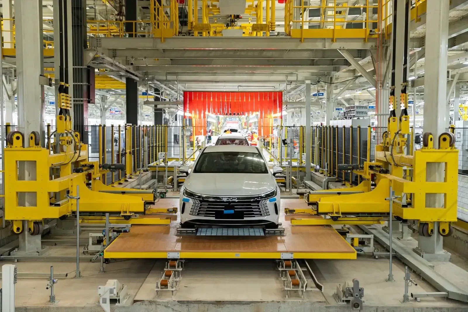 Um carro branco sendo montado em uma linha de produção industrial, com máquinas amarelas e esteiras transportadoras. Ao fundo, uma cortina vermelha e outros carros em processo de montagem